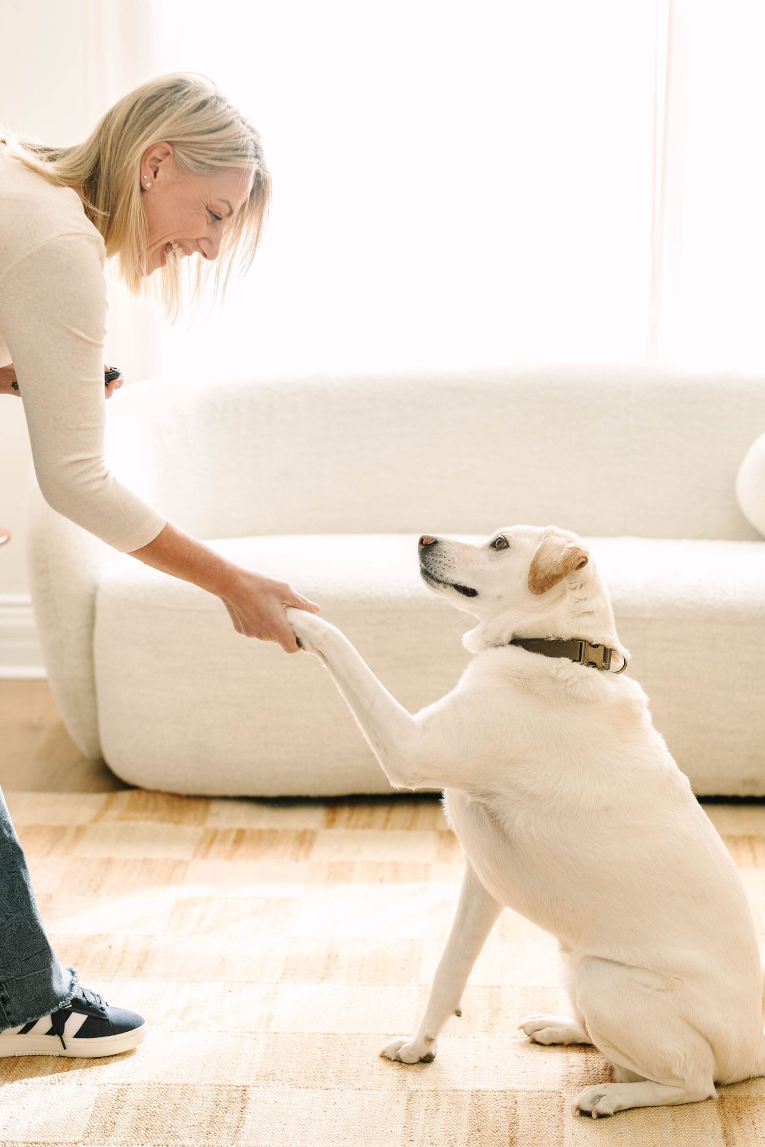 A person in white clothing sits on a light colored floor playing with a white dog in a bright sunlit room.
