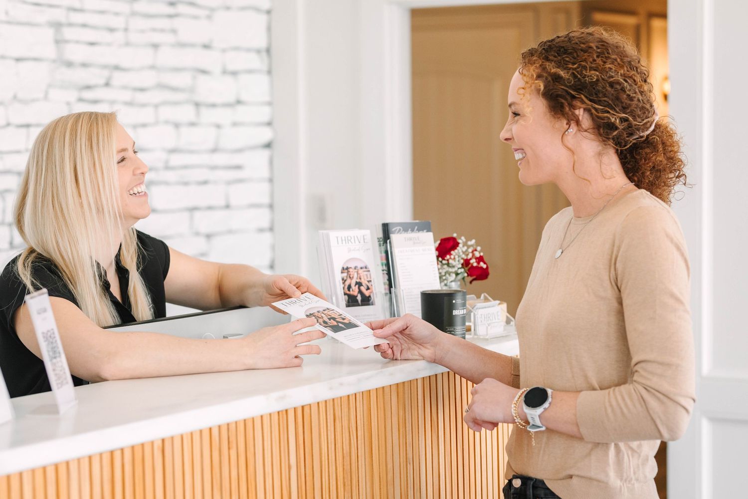 Two women smiling at a modern reception desk, one handing over a card in a bright, welcoming salon or spa.
