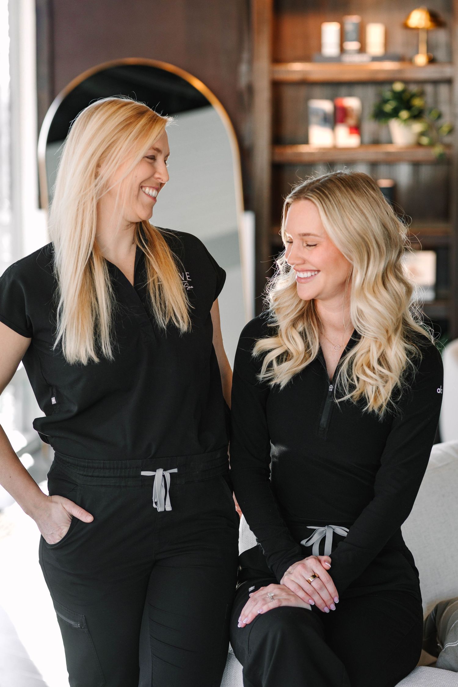 Two blonde women in black scrubs smiling together in a modern medical spa interior.
