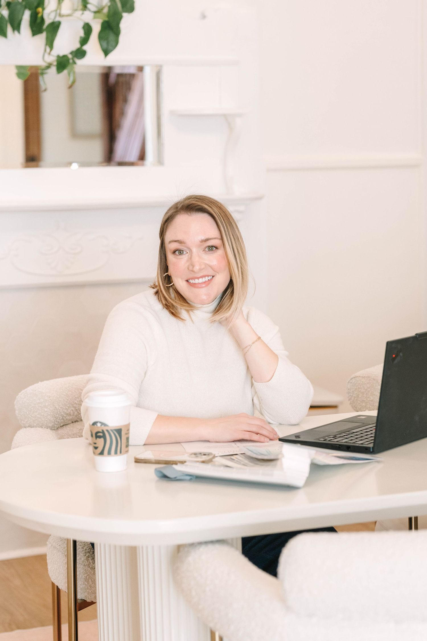 Remote worker in cozy white sweater sits at modern home office desk with laptop and coffee.