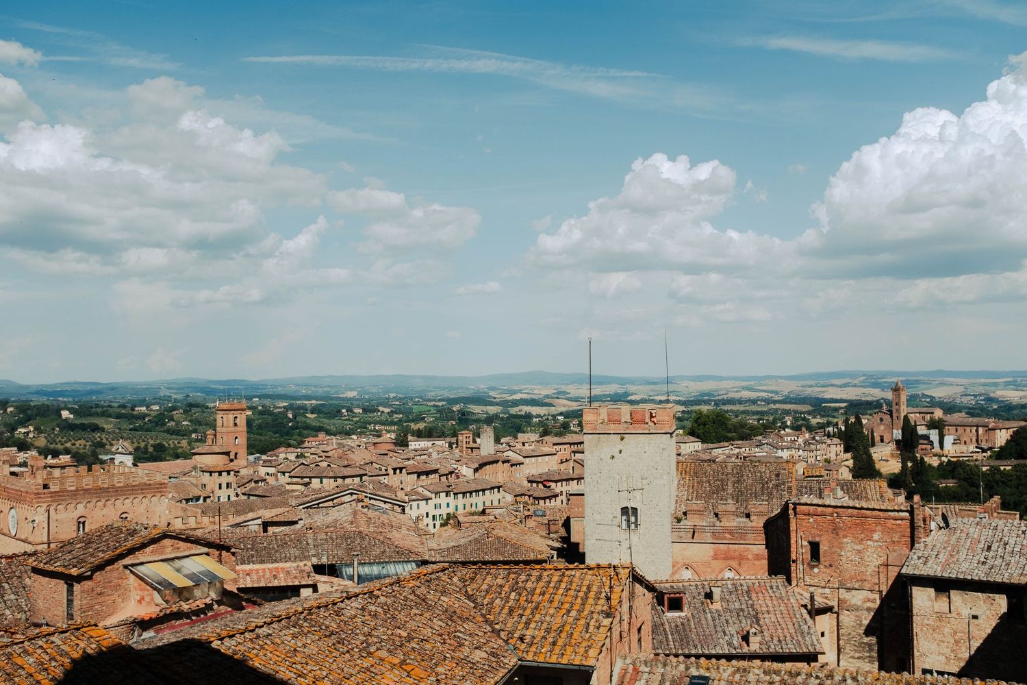 Panoramic rooftop view of a historic Tuscan hilltop town with terracotta roofs and rolling countryside under a cloudy sky.