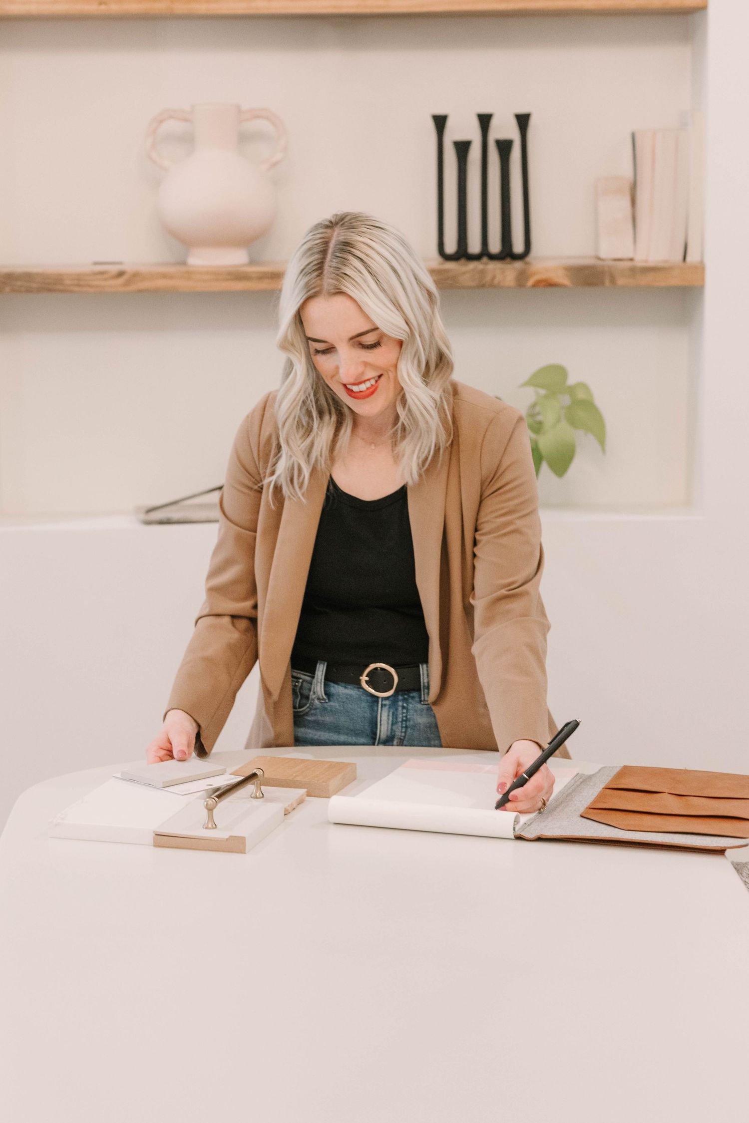 Person in tan blazer writing at a clean white desk with wooden shelving in background.
