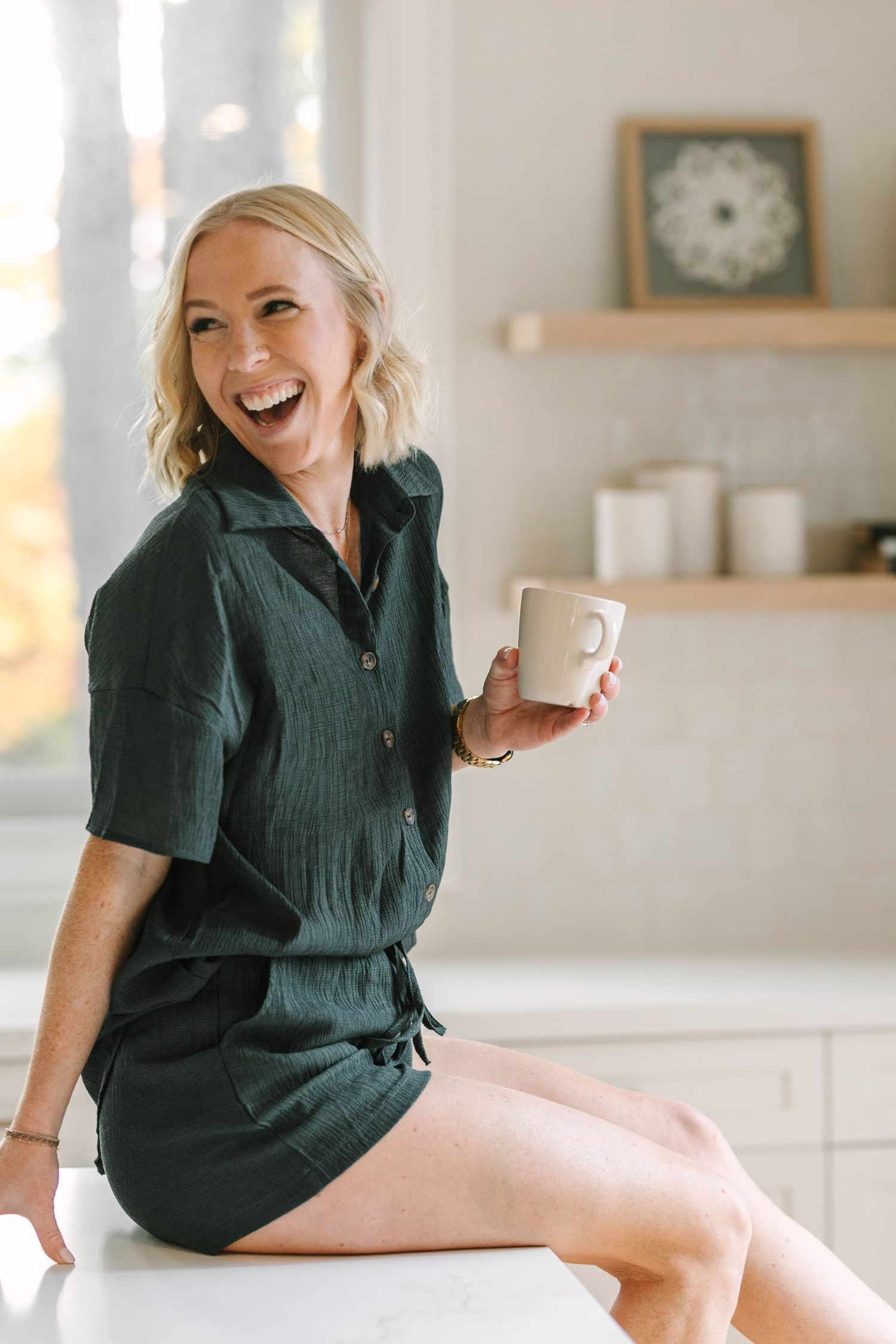 Smiling blonde woman in dark green romper sitting on kitchen counter holding a white coffee mug.