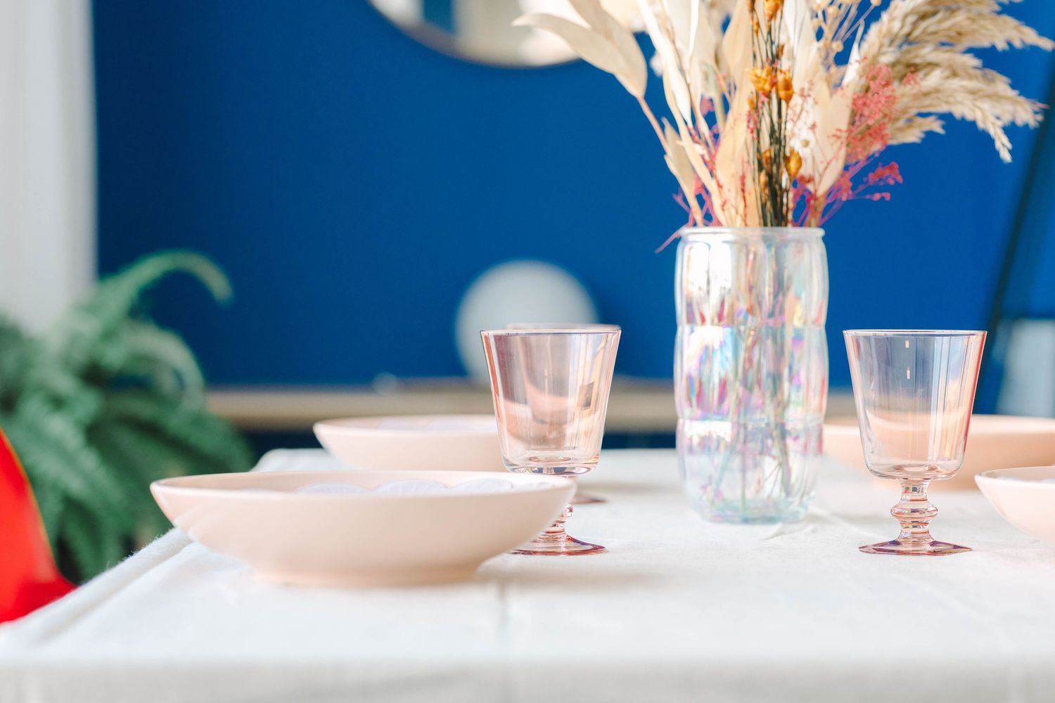 Elegant table setting with pink glassware, ceramic bowl, and dried floral arrangement in iridescent vase on white tablecloth.