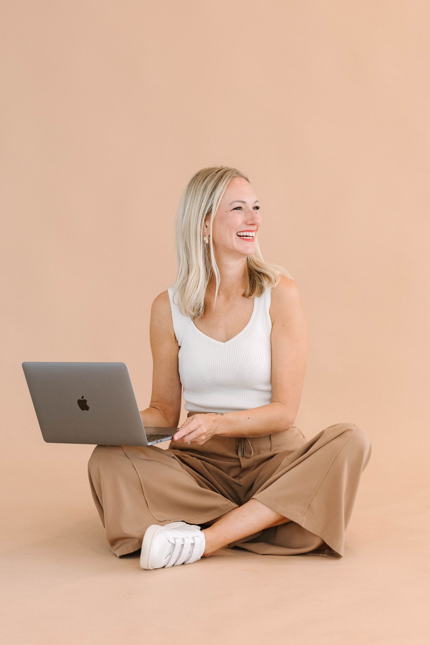 Person in casual attire sitting cross-legged with laptop on beige background.