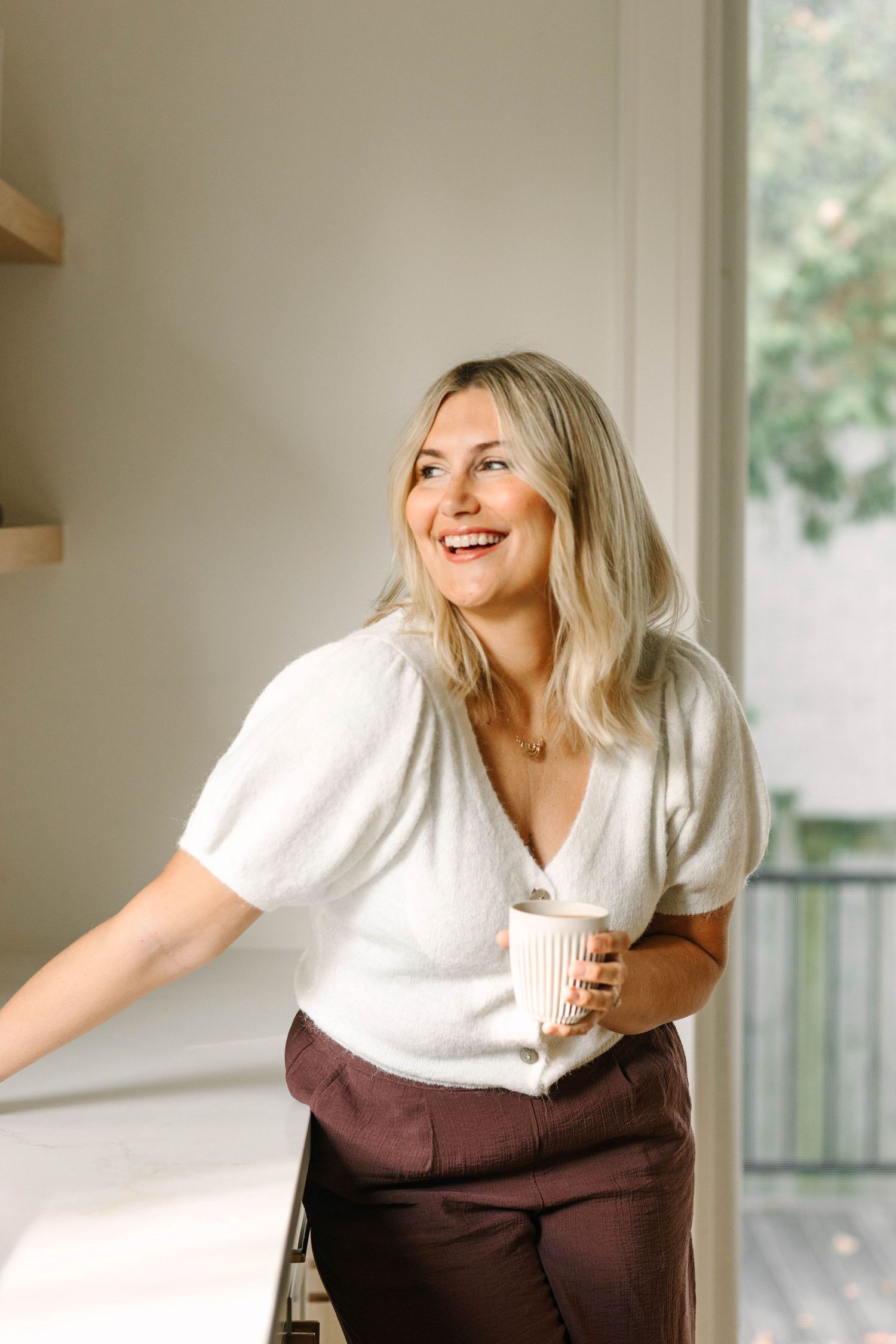 Smiling blonde woman in white top and burgundy pants holding a coffee cup near a bright window.