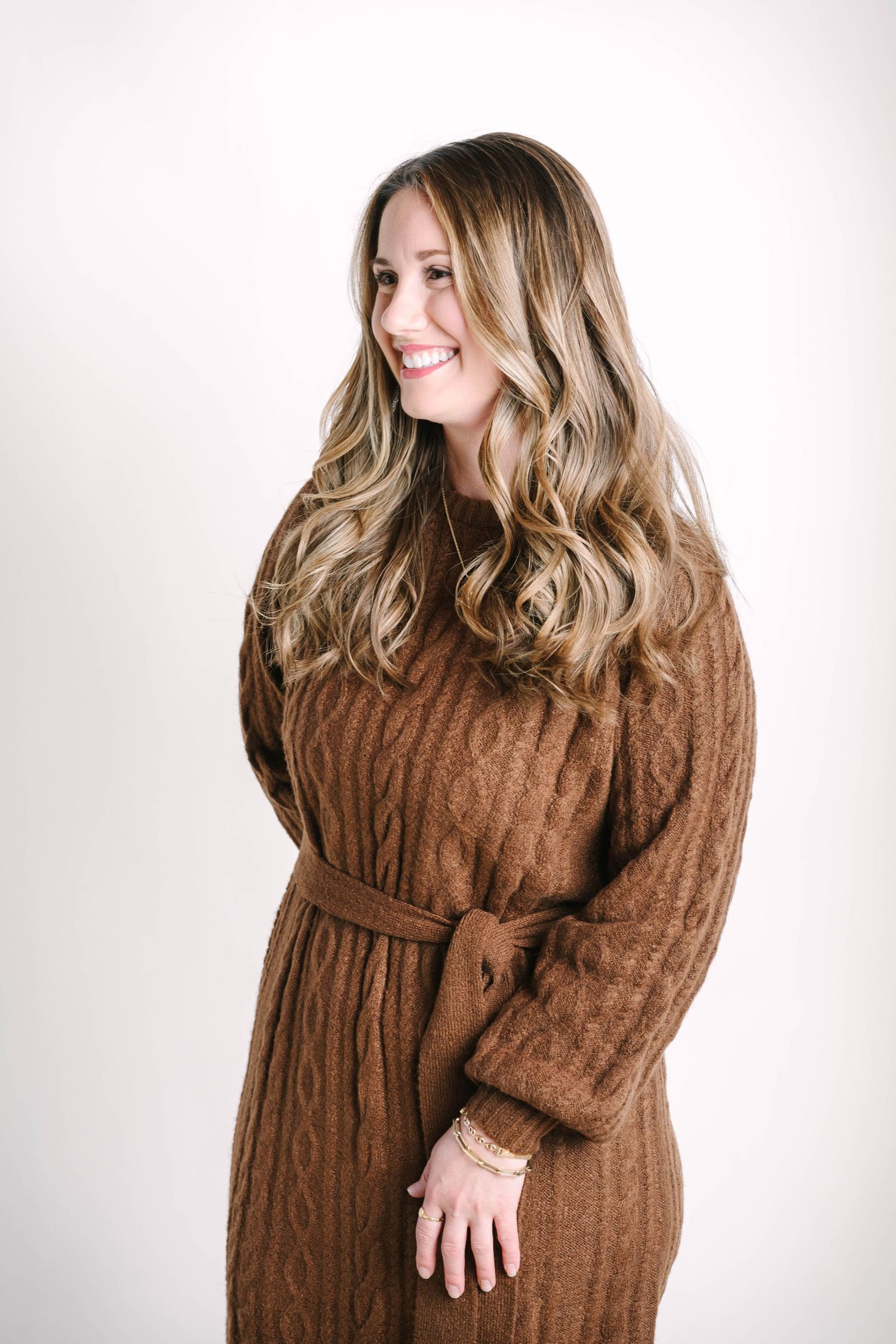 Woman in brown sweater dress poses against white background showing maternity style.