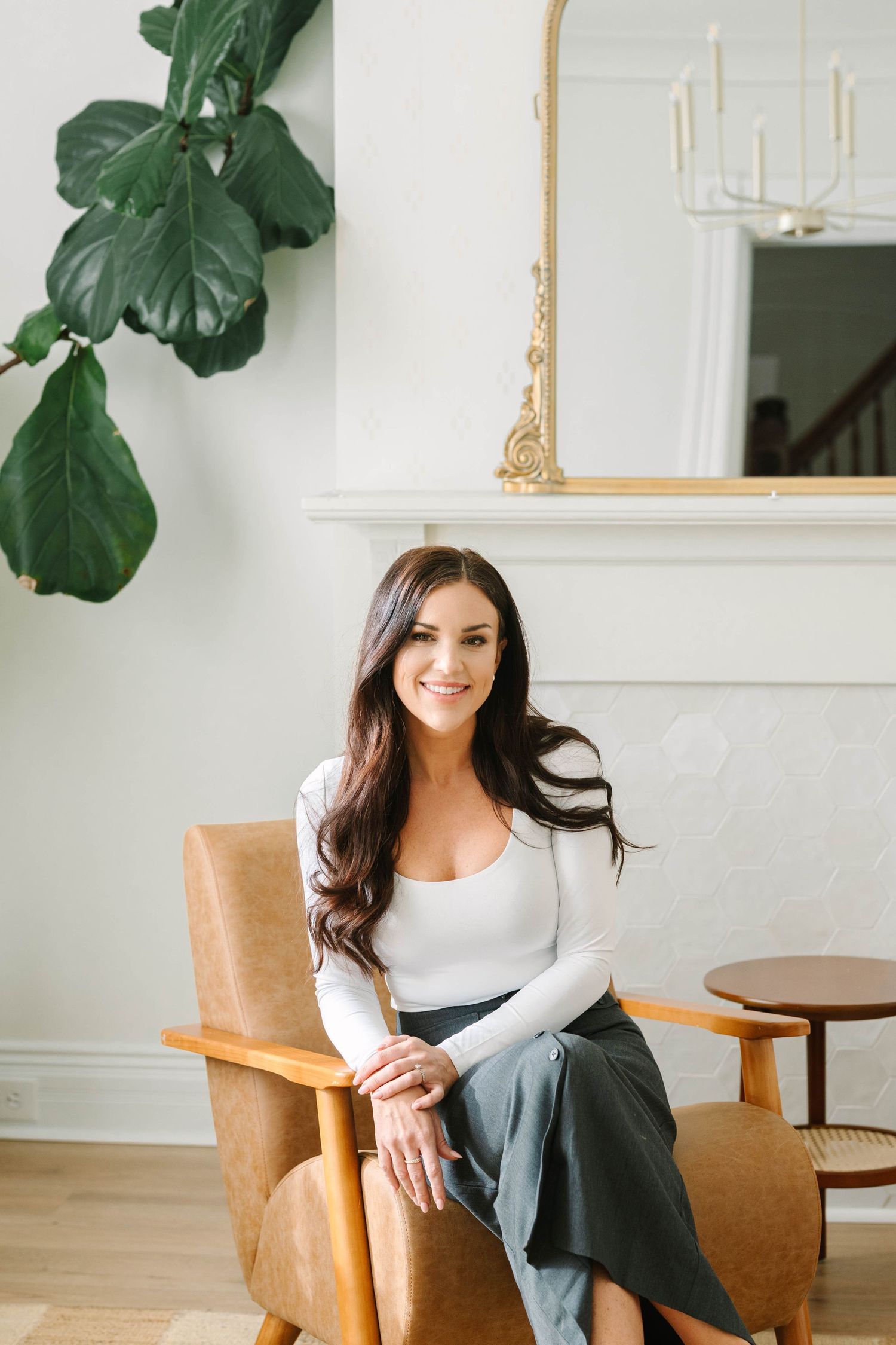 Woman in white long sleeve top and dark pants sits casually in a wooden armchair near green plants and white walls.