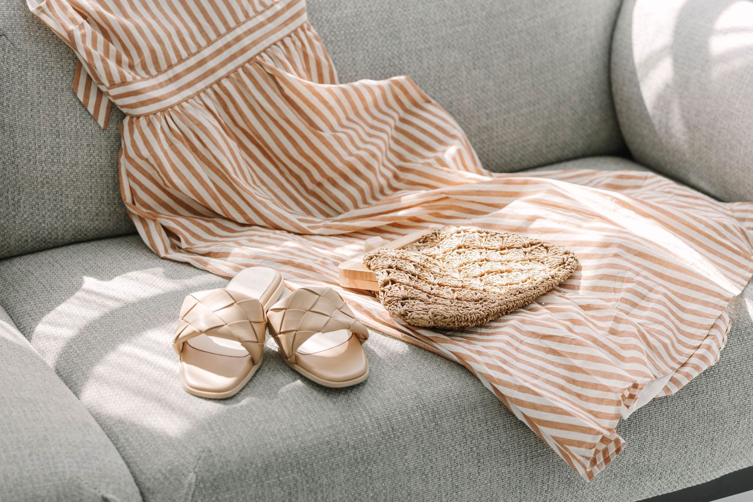 Striped beige dress, woven straw bag, and tan sandals arranged on a light gray sofa in natural sunlight.