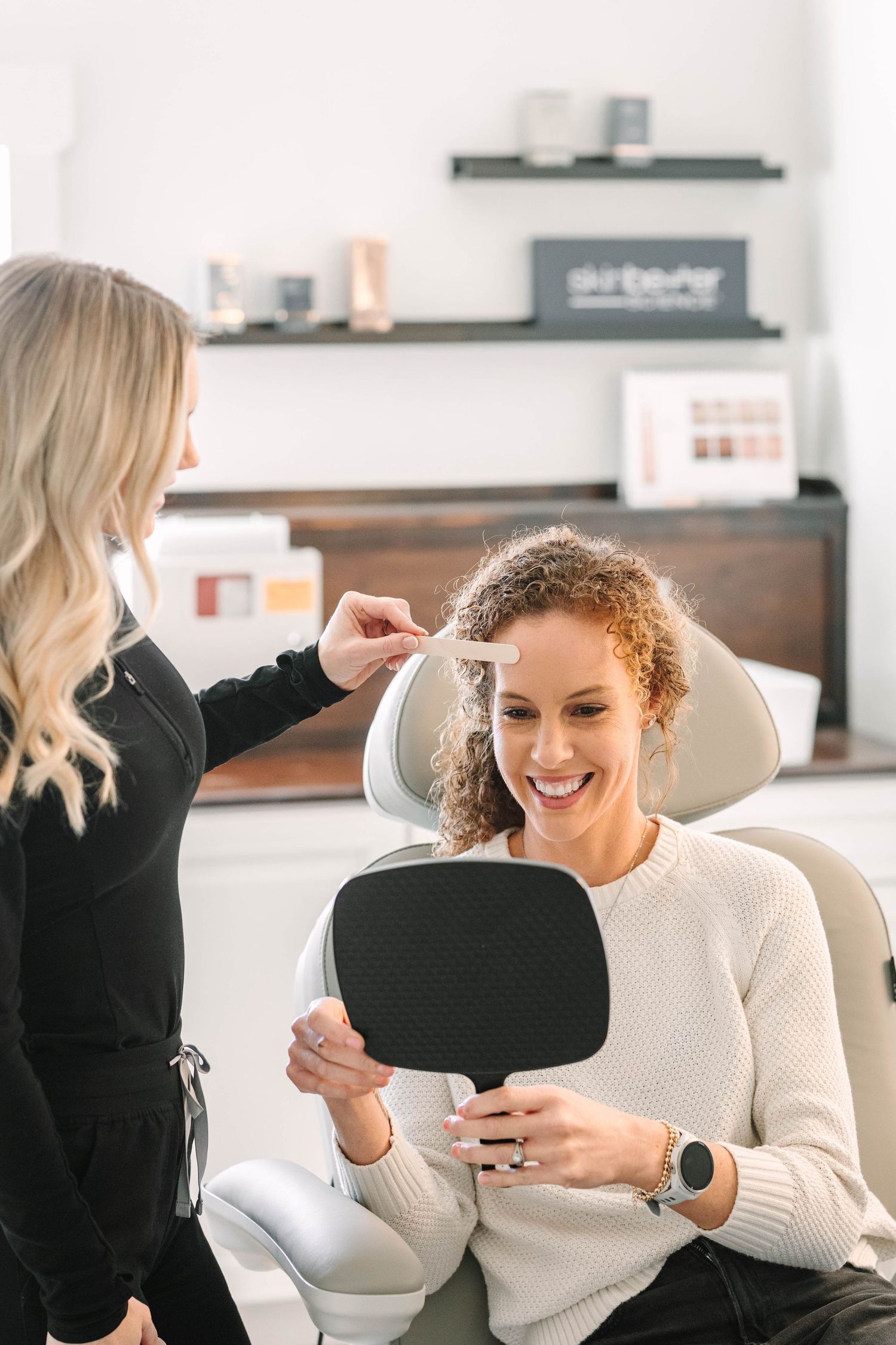 Dentist showing tooth shade sample to smiling female patient holding mirror in modern dental office.