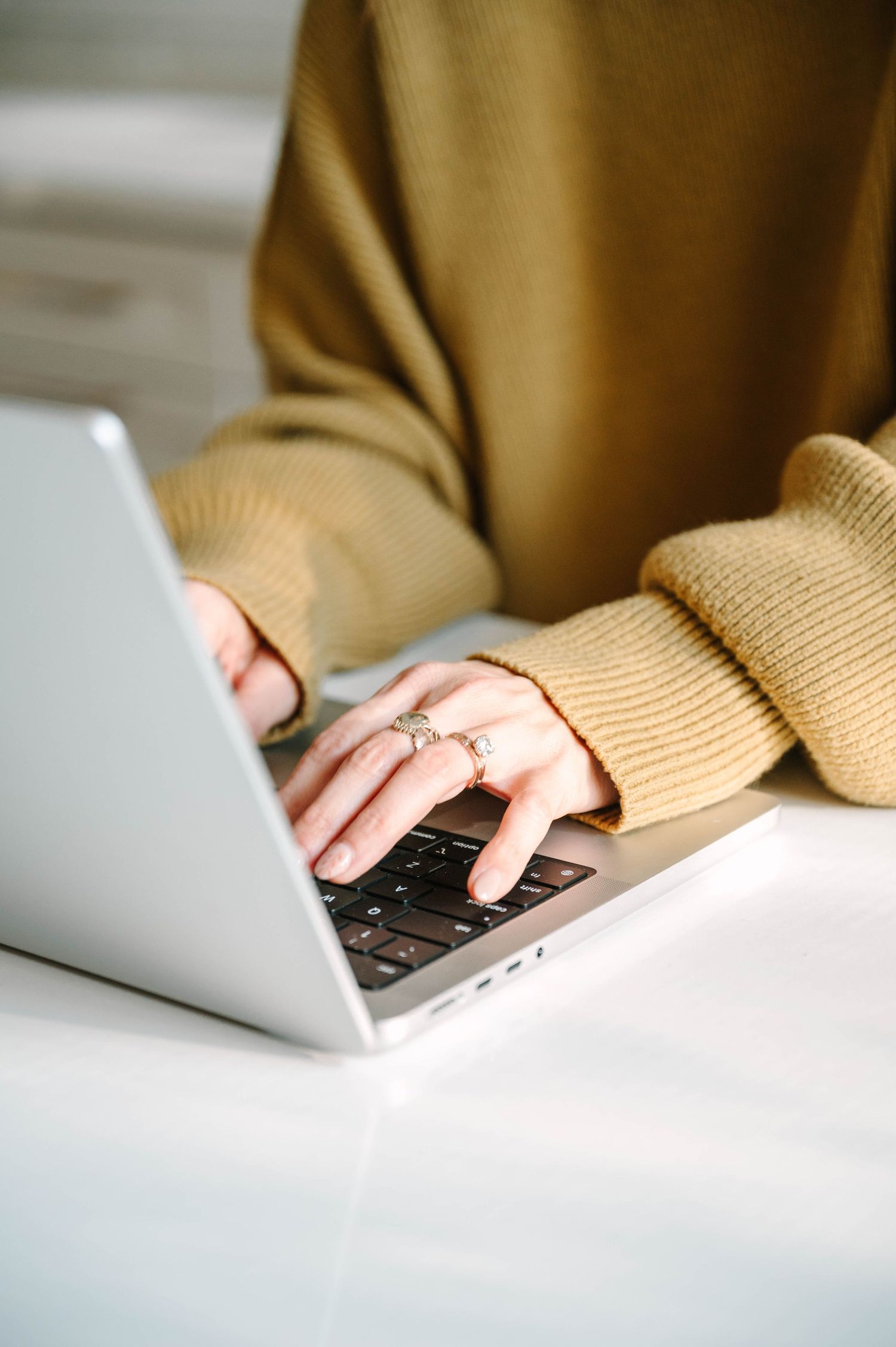 Woman in mustard sweater typing on silver laptop at white desk, wearing gold rings.