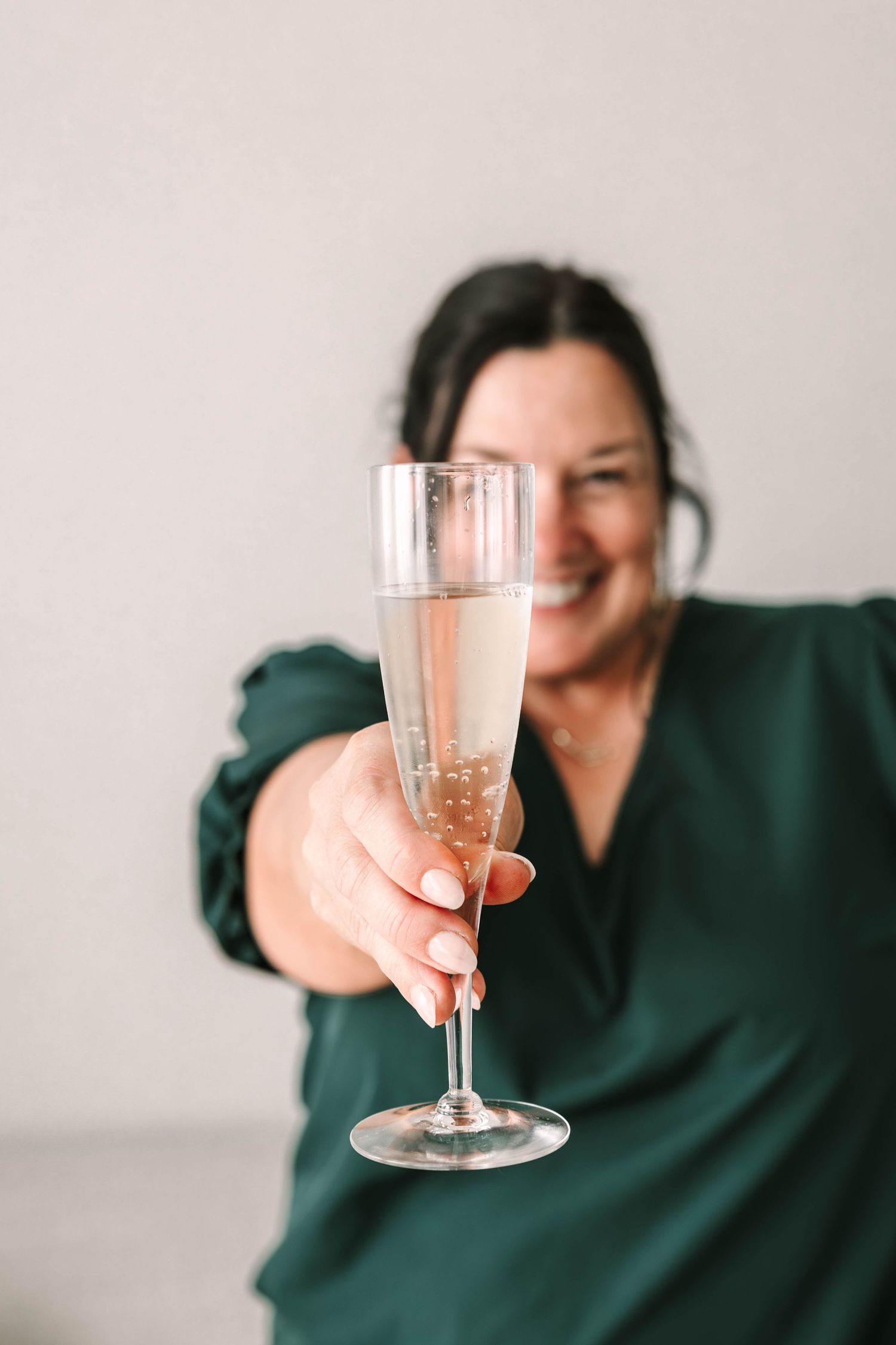 Someone in a dark green shirt holds up a glass of sparkling wine against a light background.