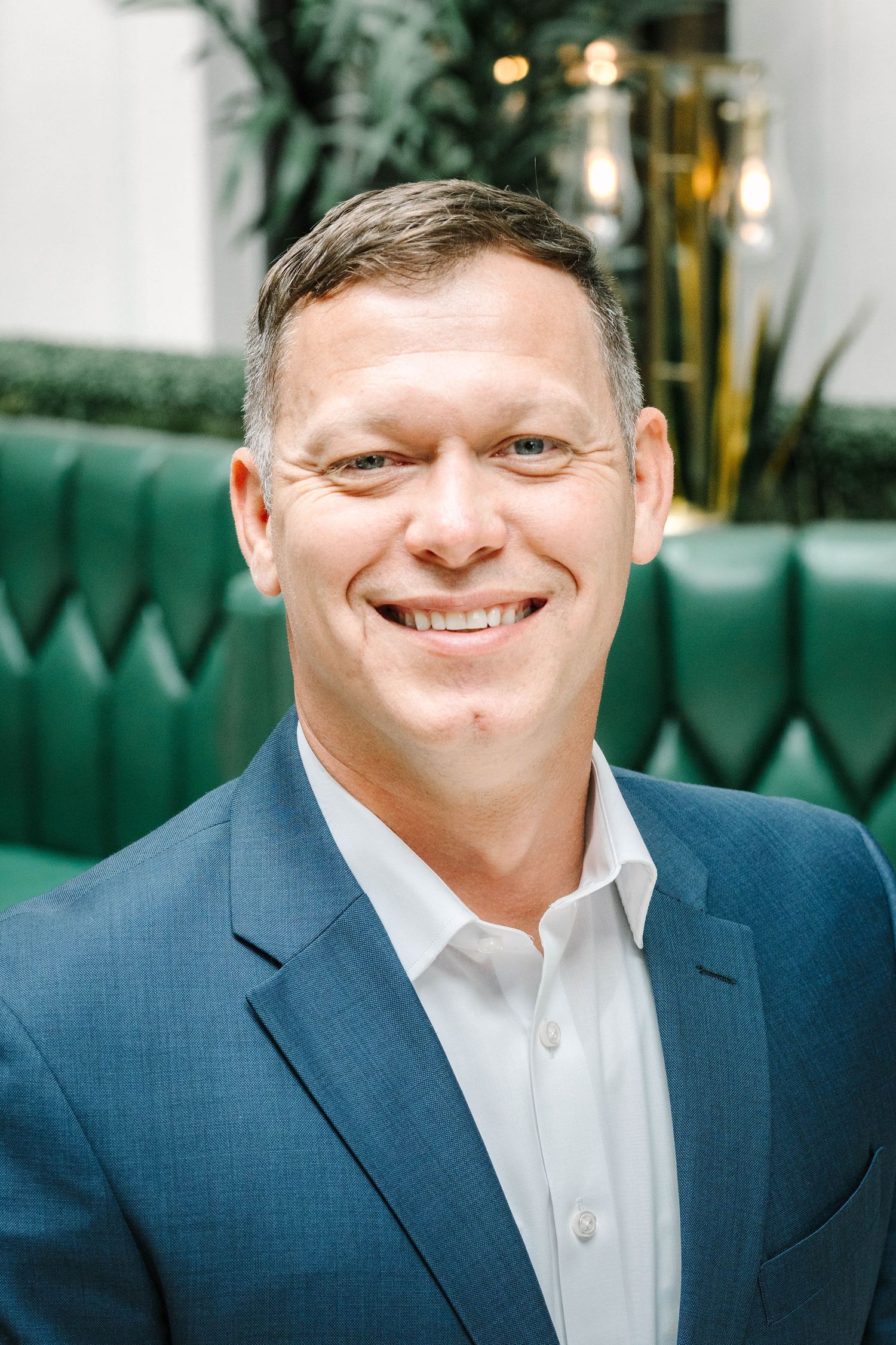 Professional headshot of person in navy suit against green tufted background.