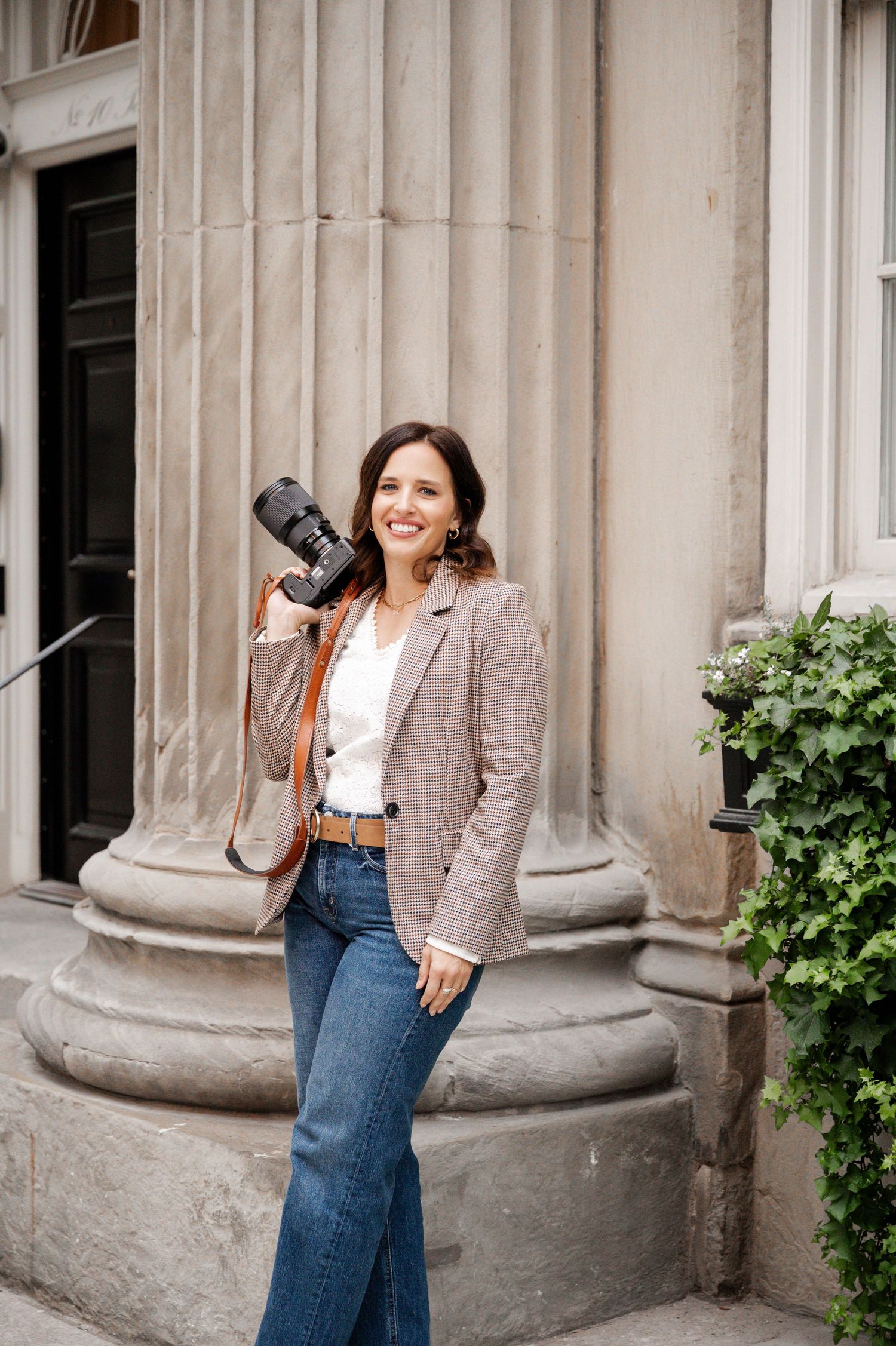 Person in blazer and jeans posing by classical stone columns with greenery.