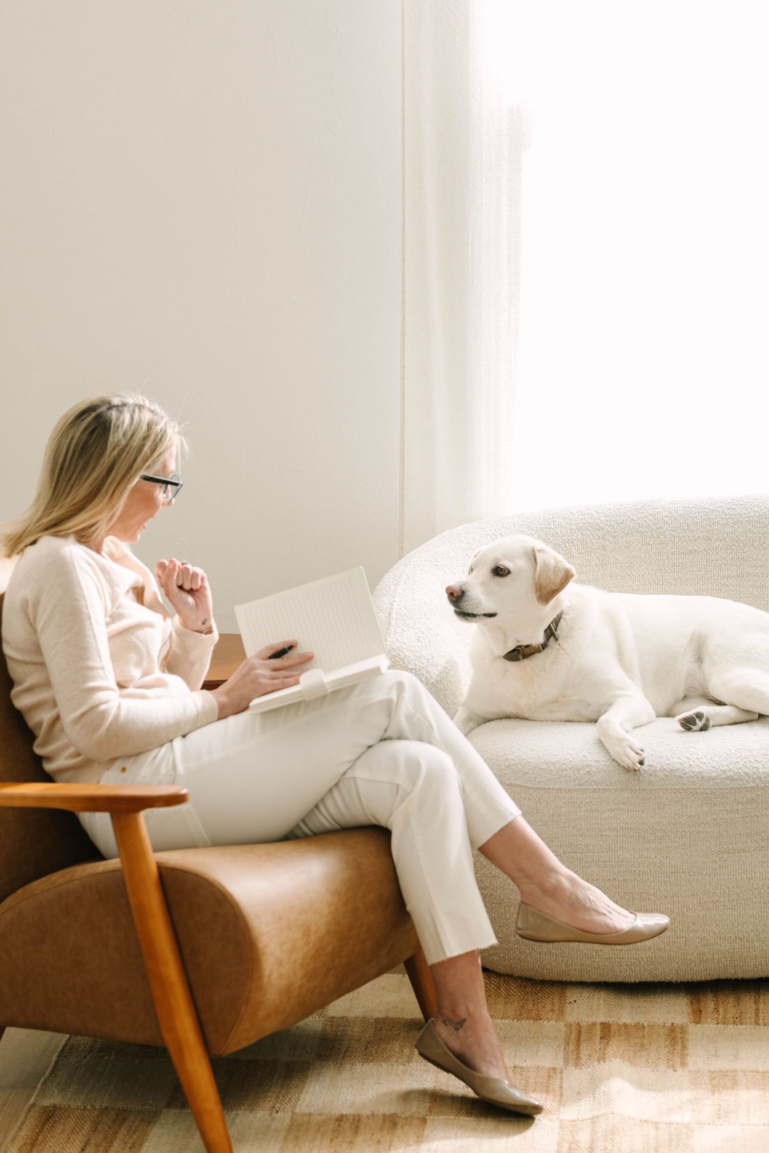 Person in white loungewear relaxes on brown leather couch while using mobile phone in bright minimalist room.