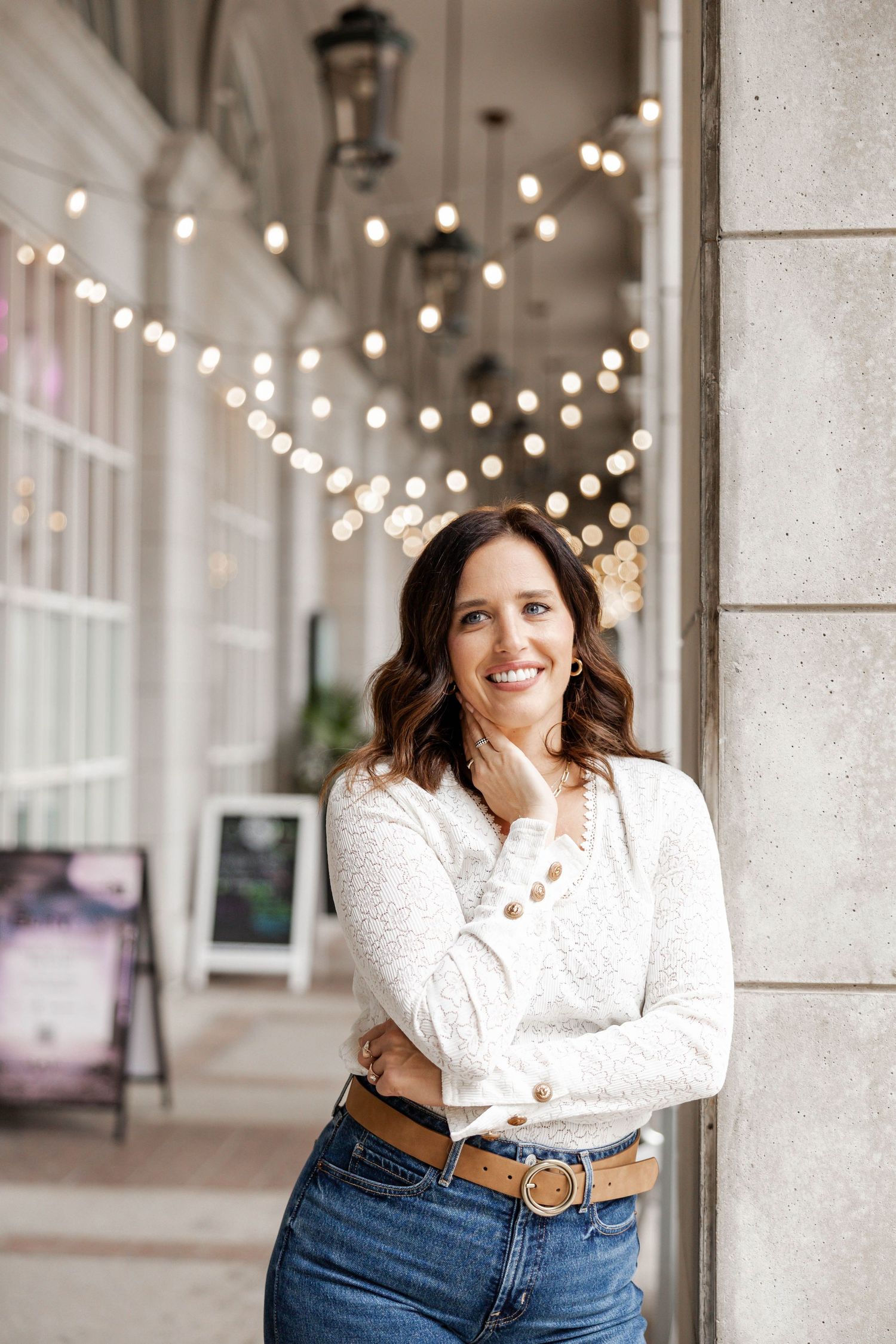 Smiling woman in white sweater and jeans leaning against column under string lights in elegant outdoor corridor.