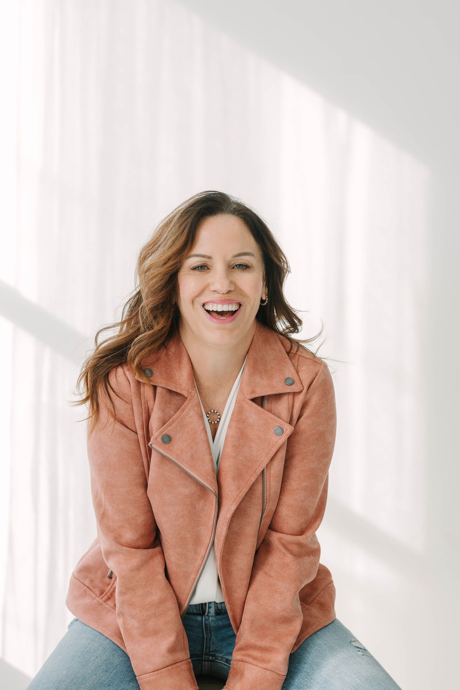 A person wearing a salmon-colored suede jacket and jeans laughs joyfully in a bright white room.