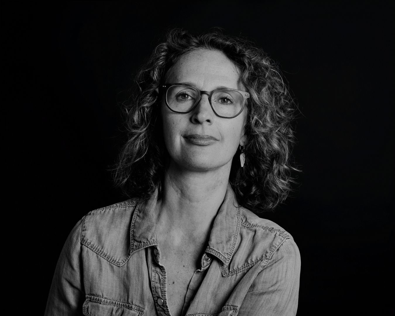Portrait featuring natural curly hair, glasses and denim shirt against dark background.