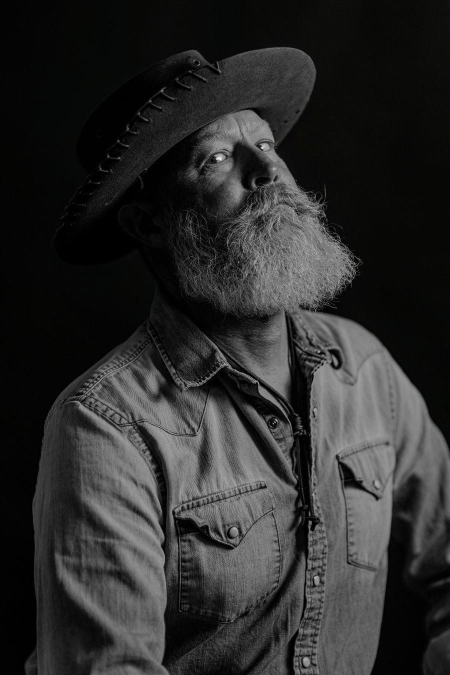 Dramatic low-key portrait showing upward gaze and full beard under wide-brimmed hat.