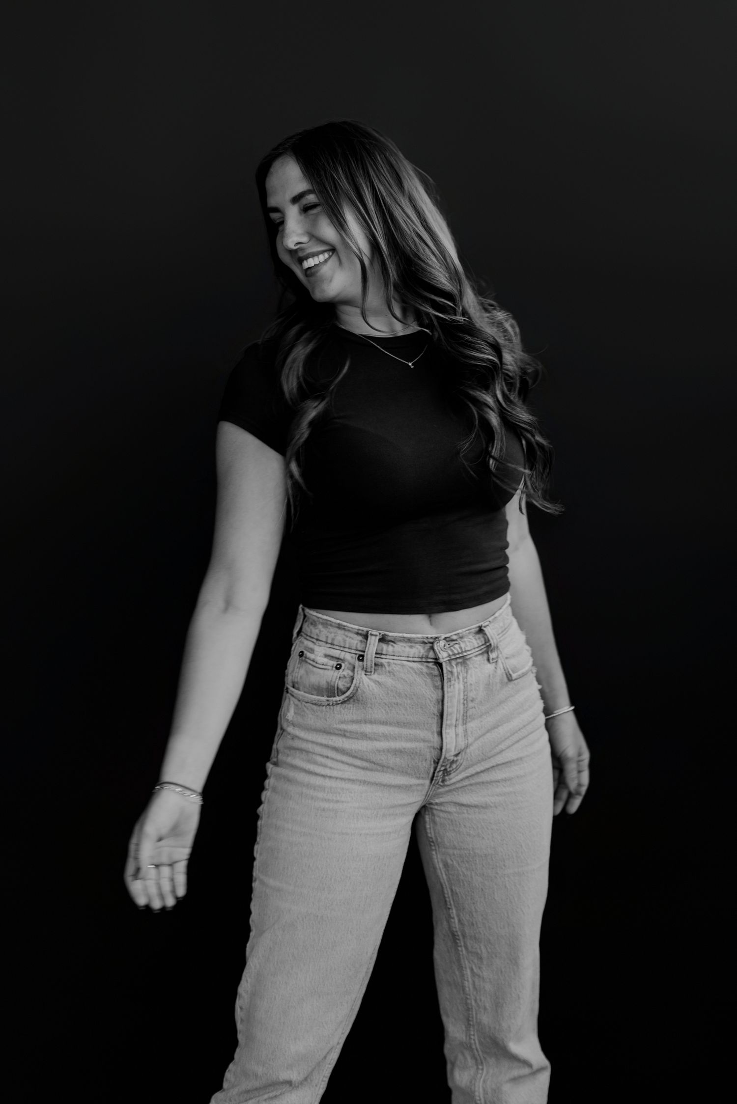 Woman in casual outfit of black top and light jeans poses against dark backdrop.