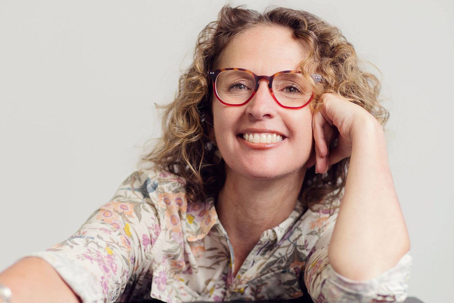 Professional headshot with lifestyle portraits showing warm smile, curly hair, and red glasses in natural lighting.