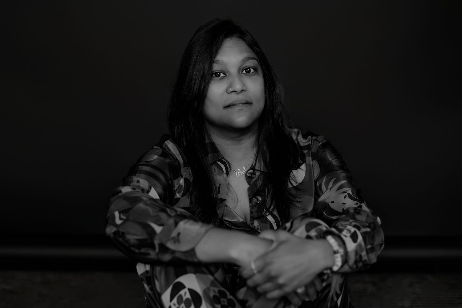 Person in dark room wearing floral print top poses expressively against black background.