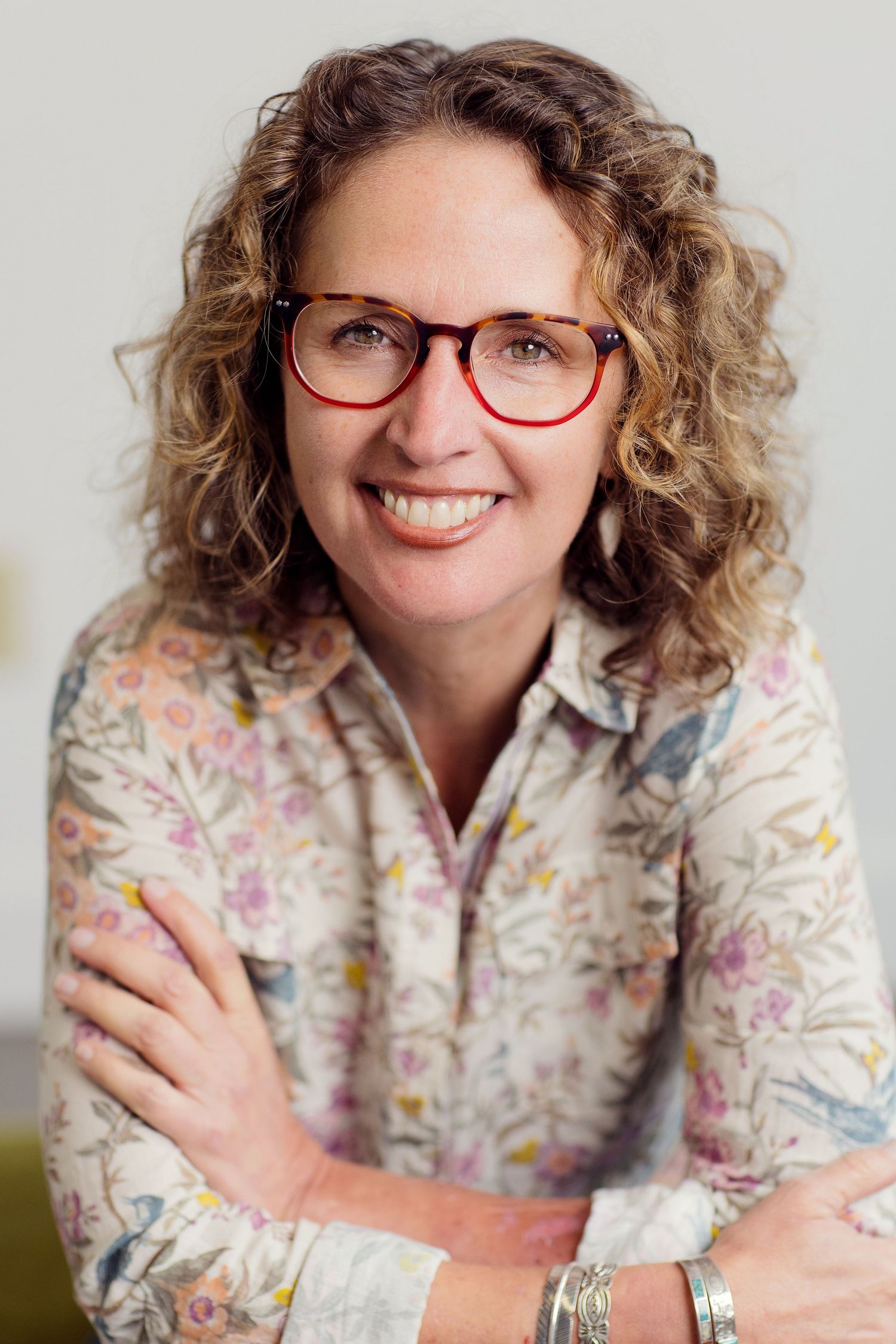 A person with curly hair and glasses wearing a floral print shirt smiles warmly at the camera in a multi-photo collage.