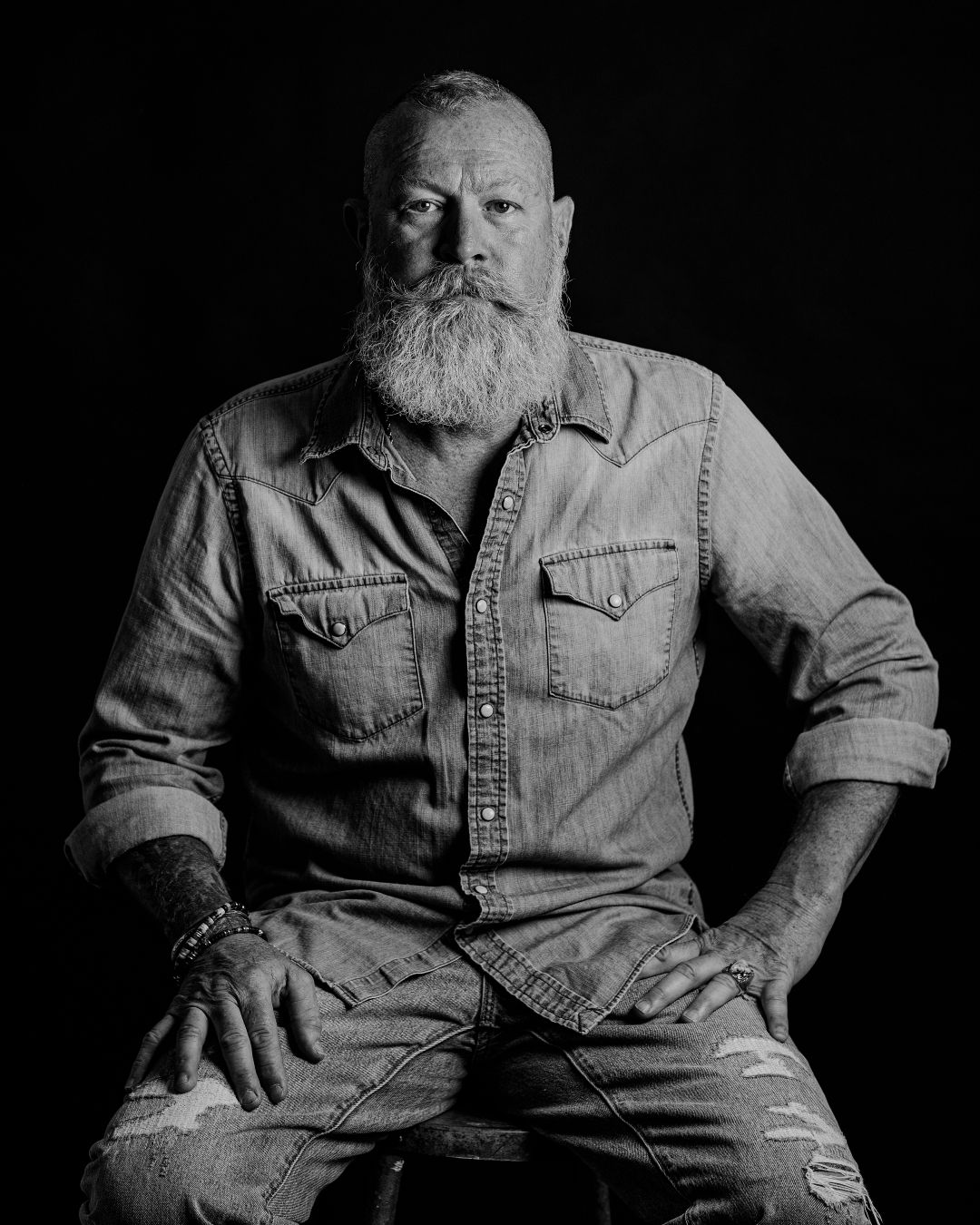 Man with full beard wearing denim shirt sits in dramatic lighting.