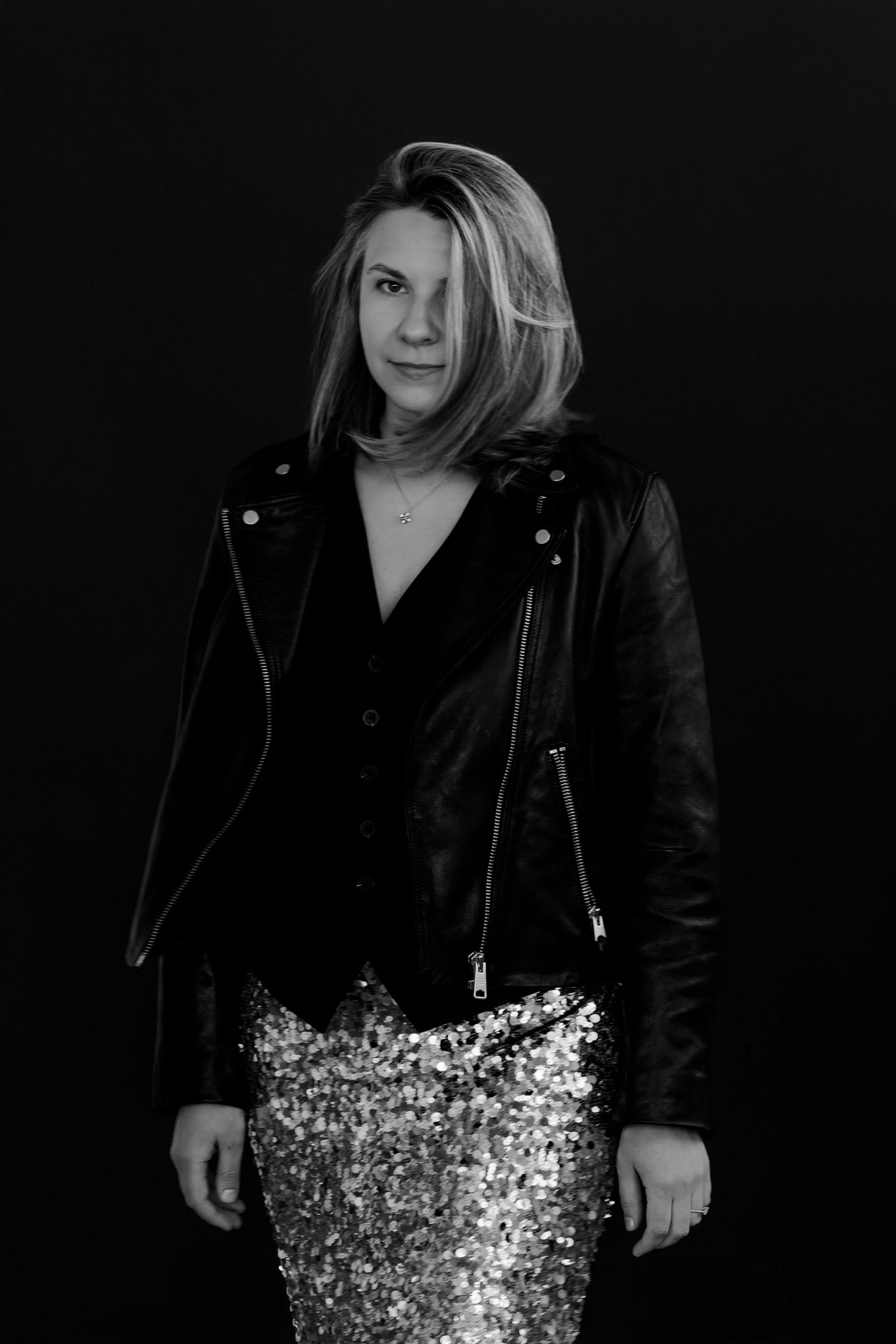 Person wearing black leather jacket and sparkly skirt poses in moody studio lighting.