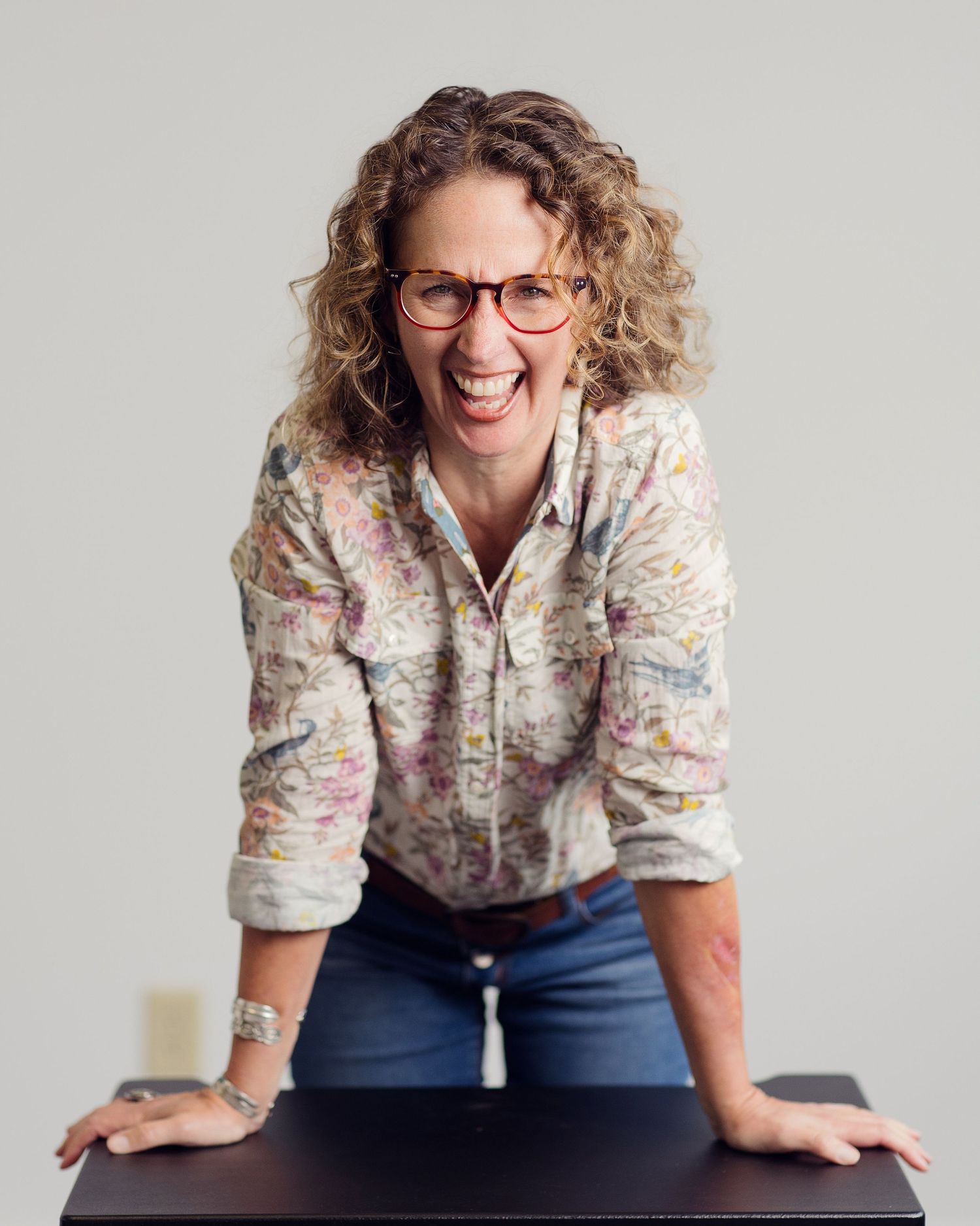 Person wearing floral print shirt and glasses sits casually on bench against light background in series of poses.