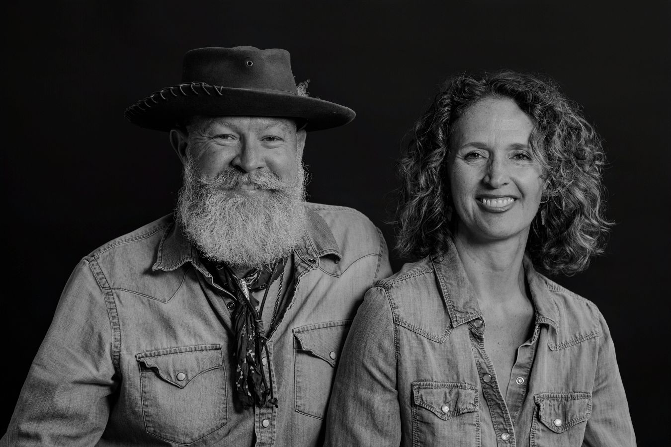 Black and white portrait of couple embracing in casual denim clothing against dark background.