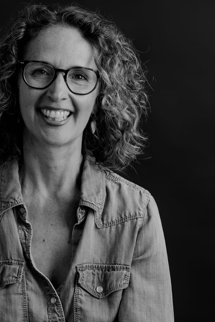 Black and white headshot of curly-haired figure in denim with glasses and bright expression.