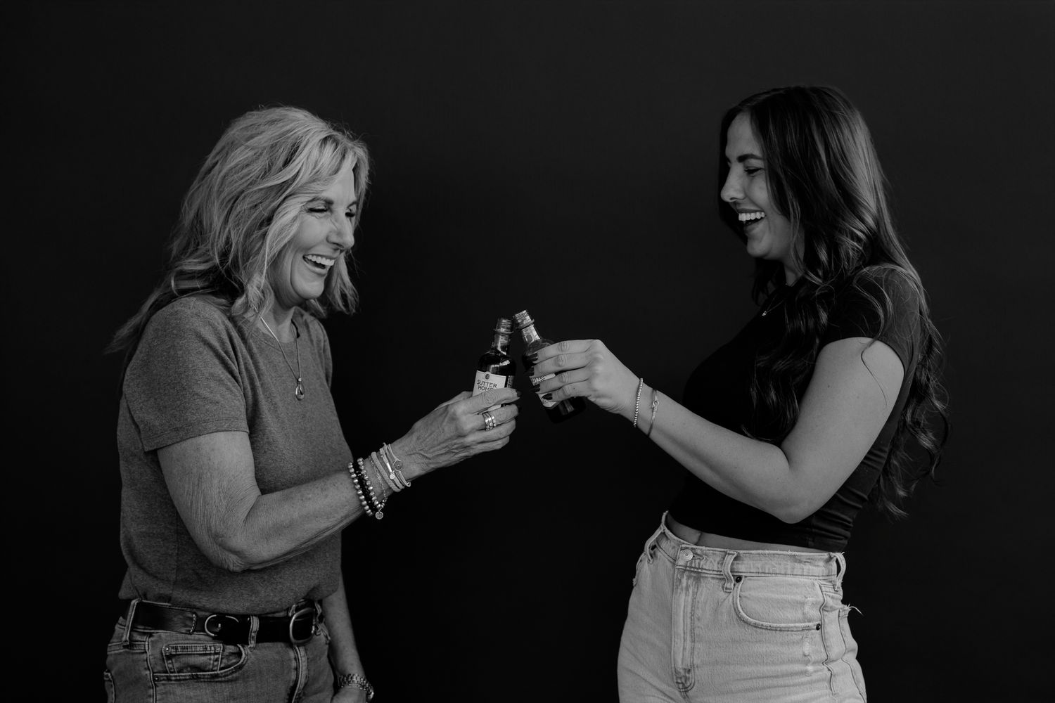 Two women laugh and smile together while sharing wine in a dark atmospheric setting.