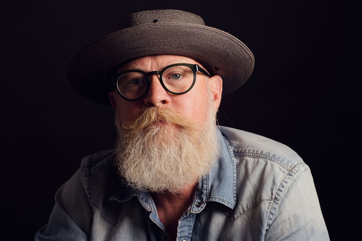 Professional studio portrait of person with gray beard wearing glasses, denim shirt and brown flat brimmed hat against dark backdrop.