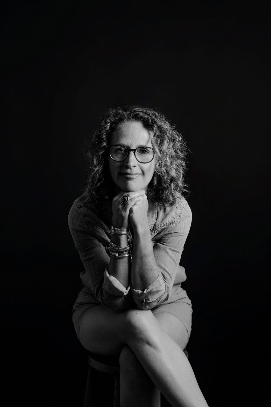 Studio portrait of seated figure in casual pose with curly hair and glasses.