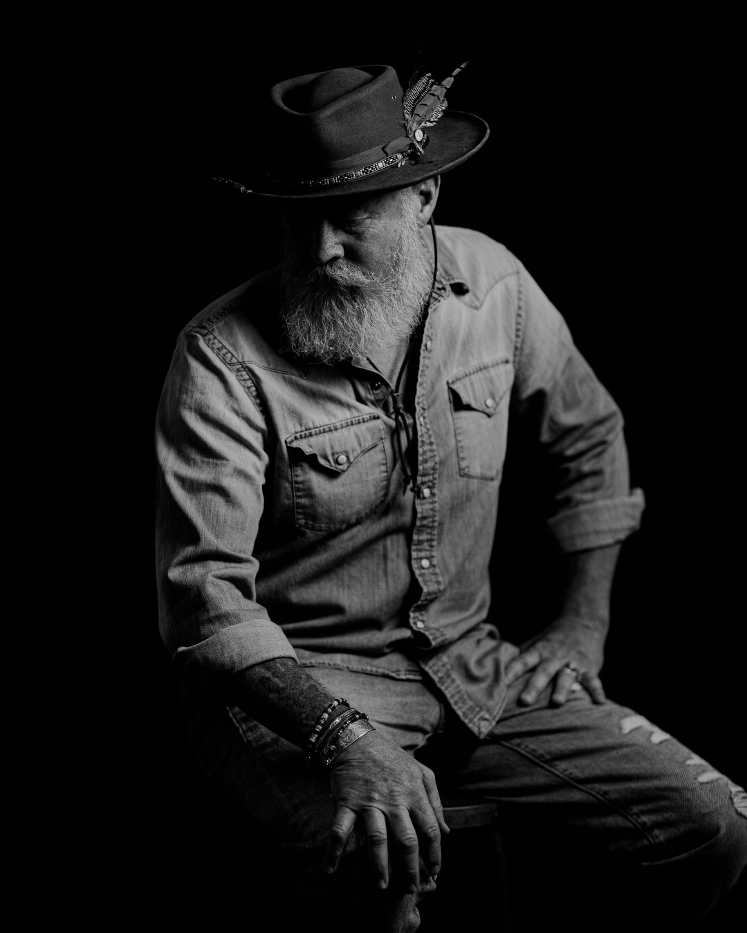 Black and white portrait of figure in denim shirt and hat sitting thoughtfully against dark background.
