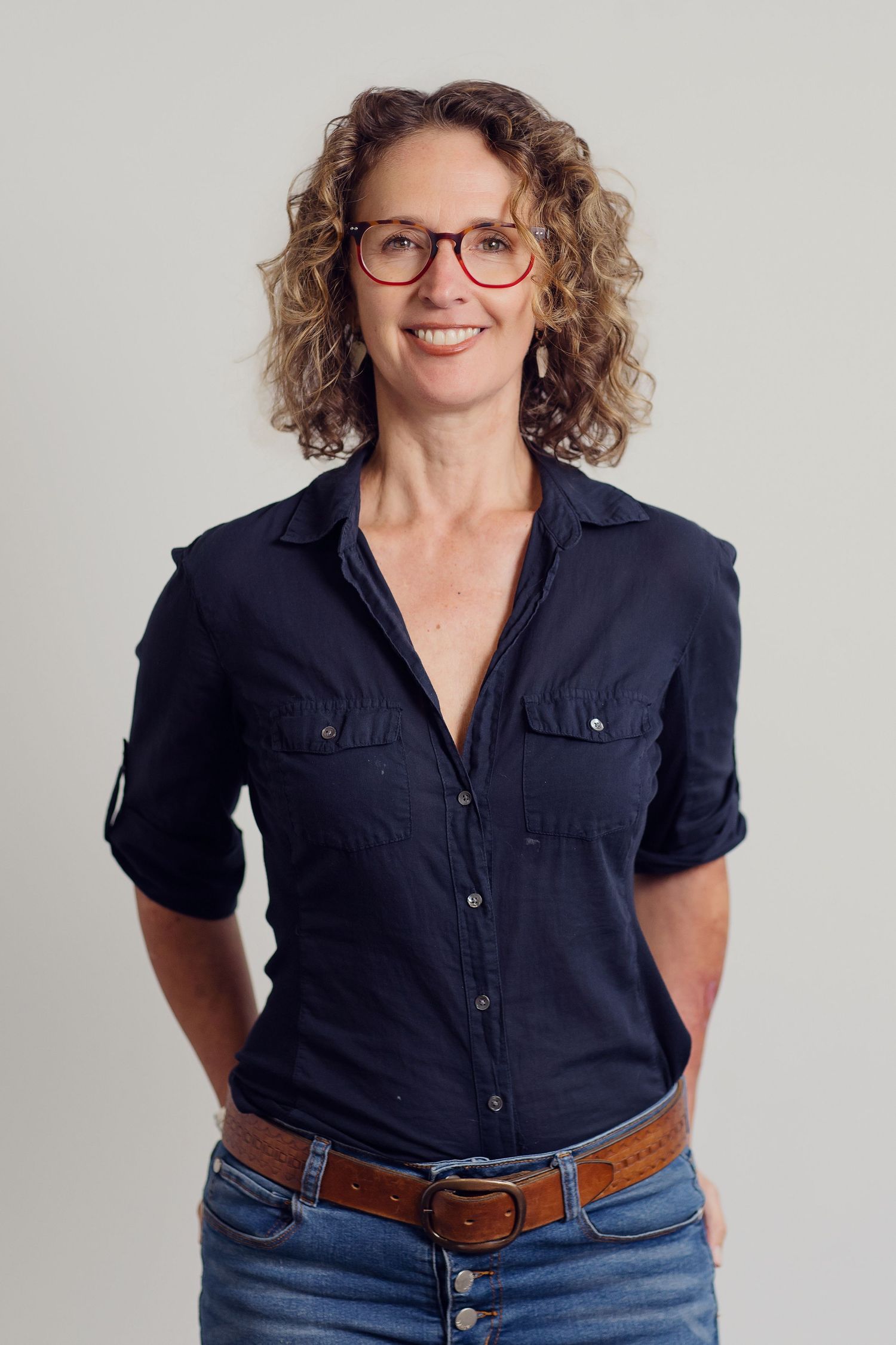 Person wearing glasses and a navy button-up shirt with curly hair poses against a light background.
