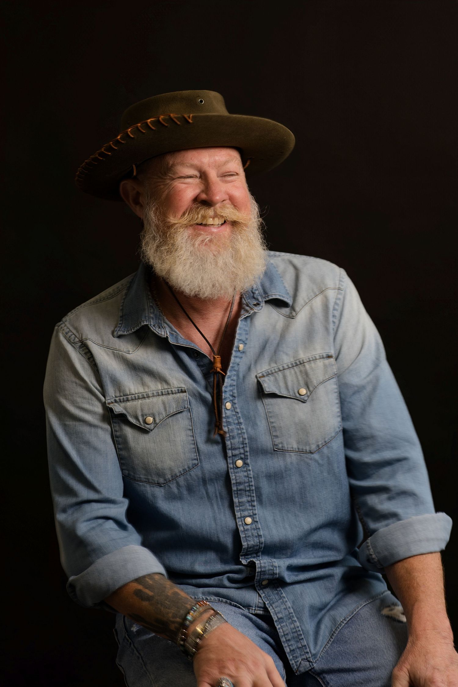 Portrait series showing an older man in denim shirt and cowboy hat against dark background, smiling and looking relaxed.
