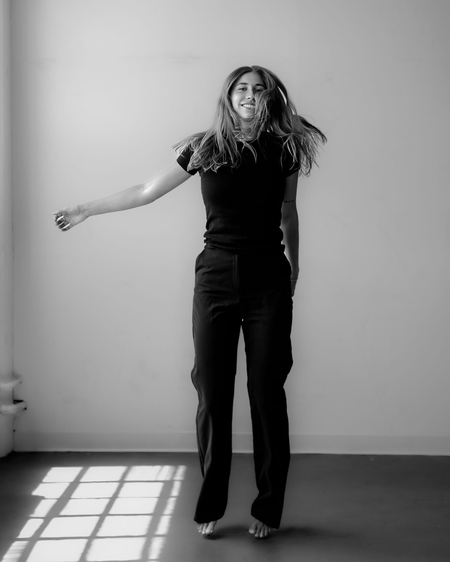 Figure in black clothing dances and moves expressively against white wall with window shadows.