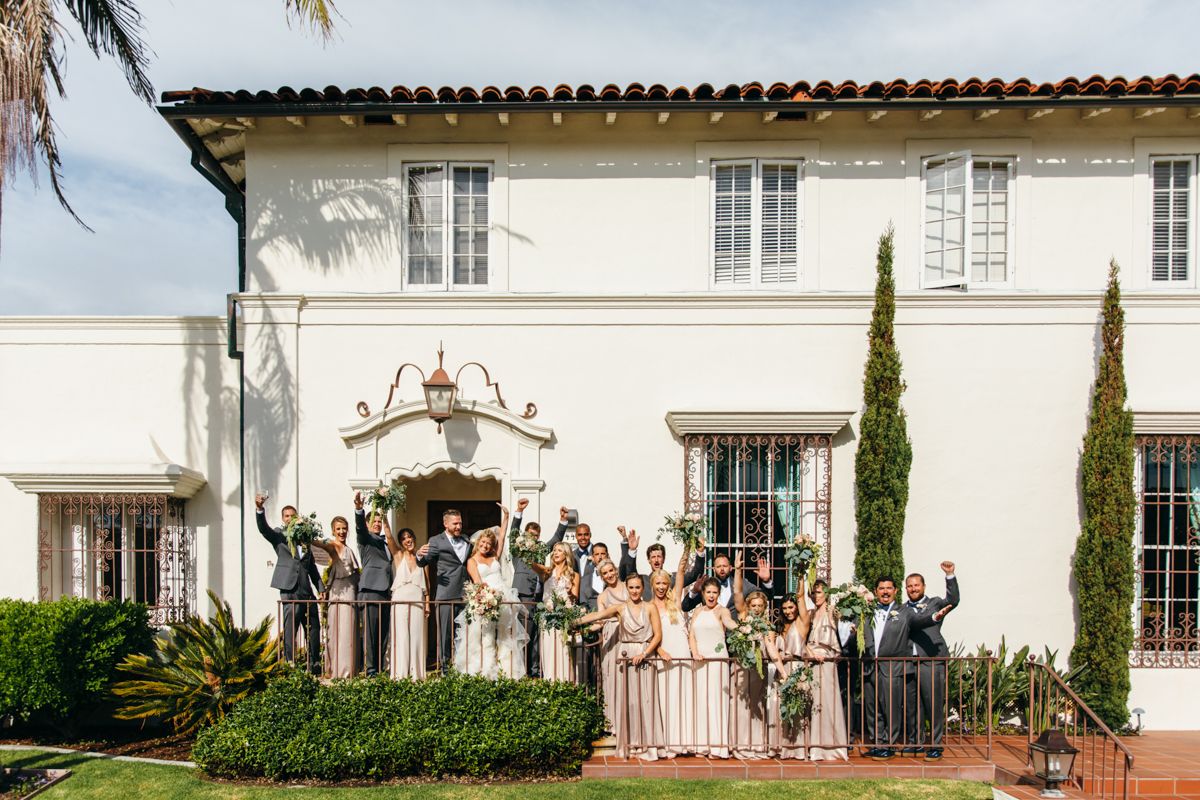 Full wedding party celebrating outside a white Spanish colonial estate on a sunny day.