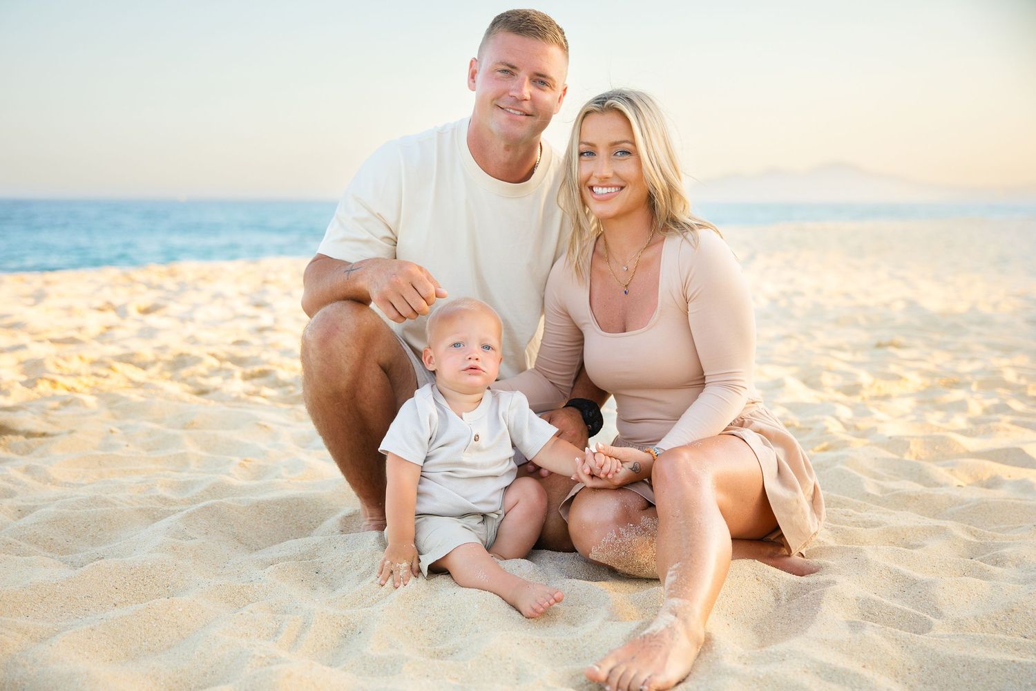 Young couple with their baby sitting together on golden sand at a sunny beach.