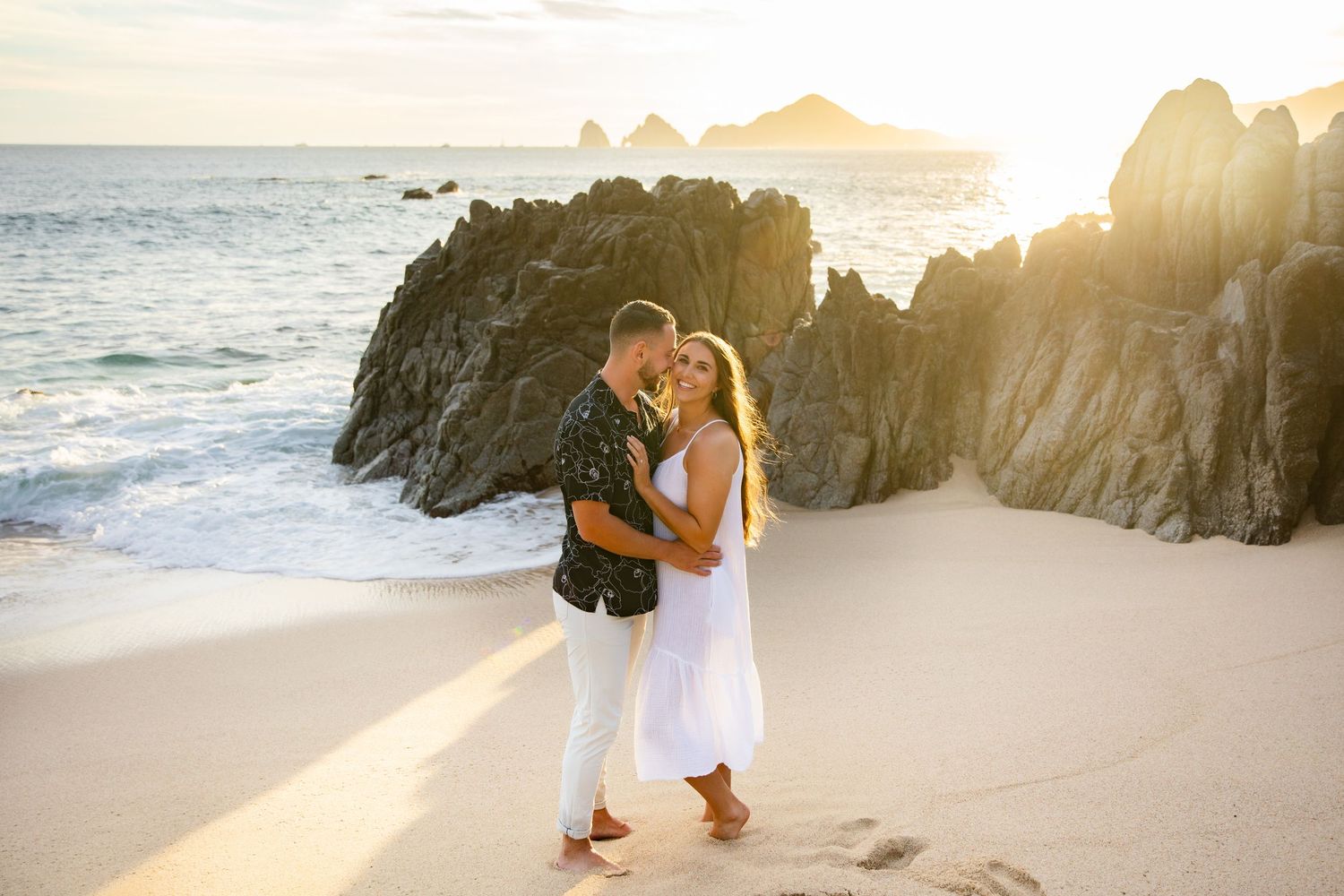 Couple sharing a romantic kiss at Playa Monuments  beach in Cabo San Lucas at golden sunset with dramatic rock formations in background.