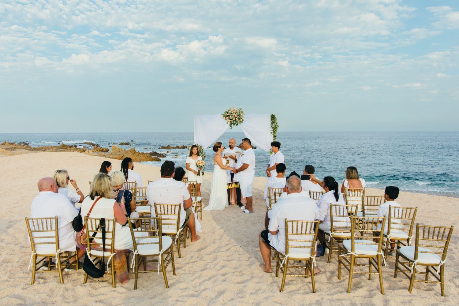 Intimate beach wedding ceremony in Cabo San Lucas with guests seated on gold chairs facing the ocean altar under a clear blue sky.