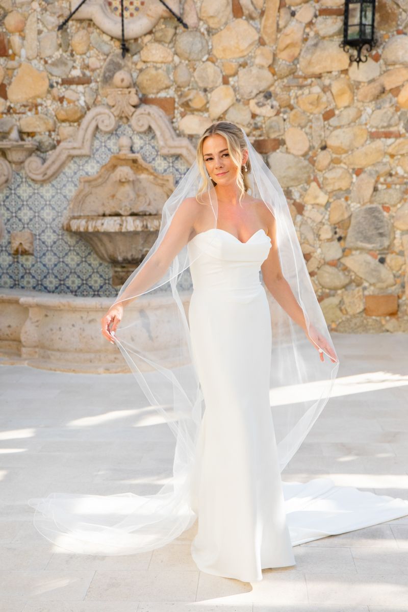 Elegant bride in strapless white gown and veil poses before rustic stone wall fountain.