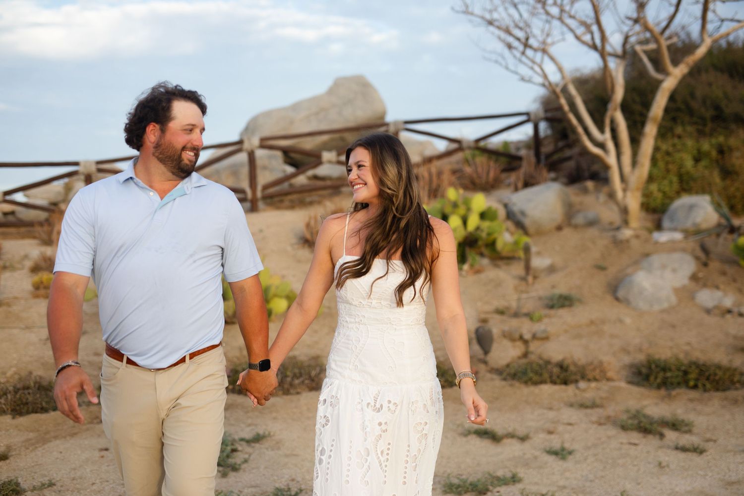 Couple holding hands and smiling while walking on a sandy desert path with rocks and cacti in the background.