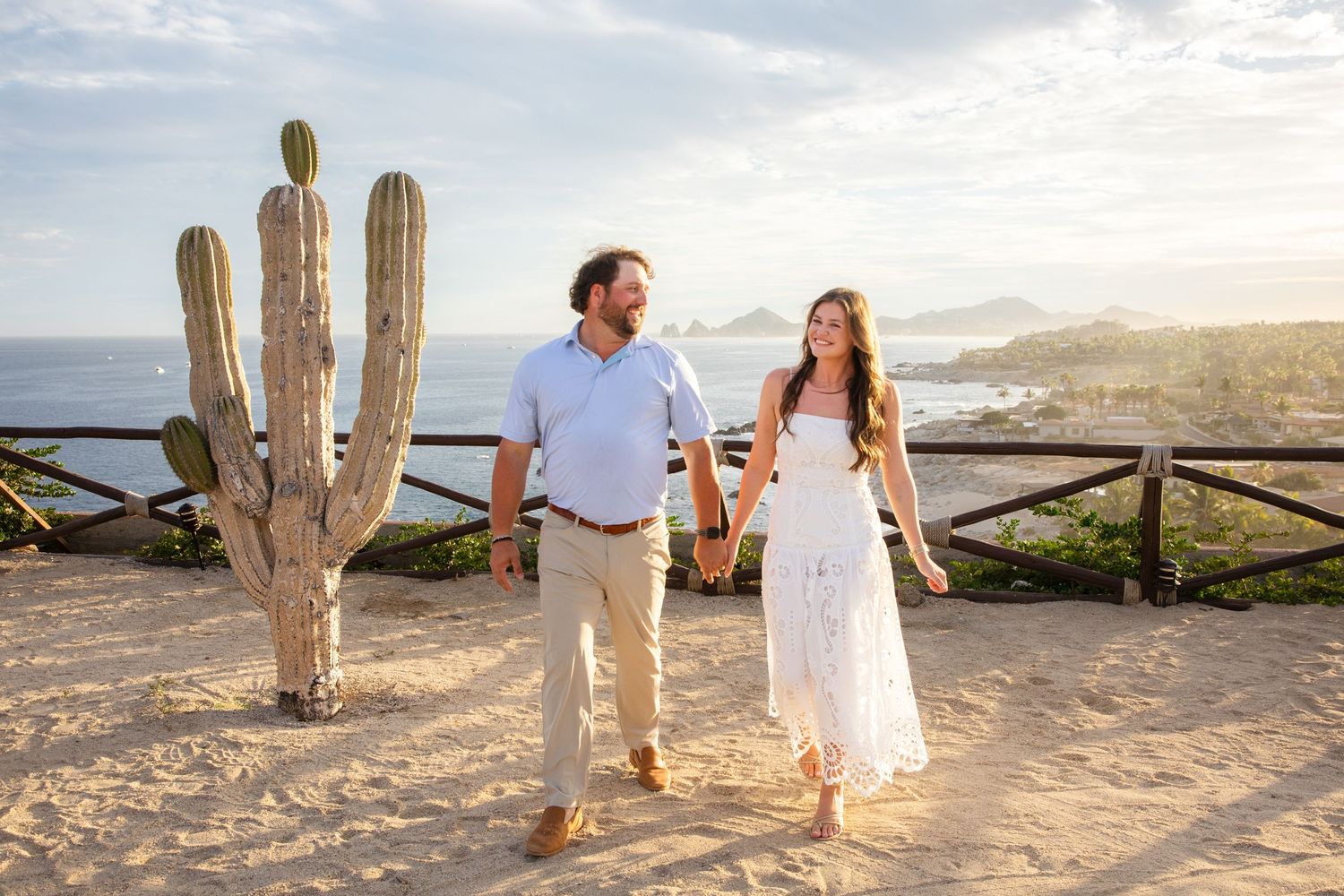 Couple holding hands walking on sandy overlook with cactus and ocean view in Los Cabos, Mexico.