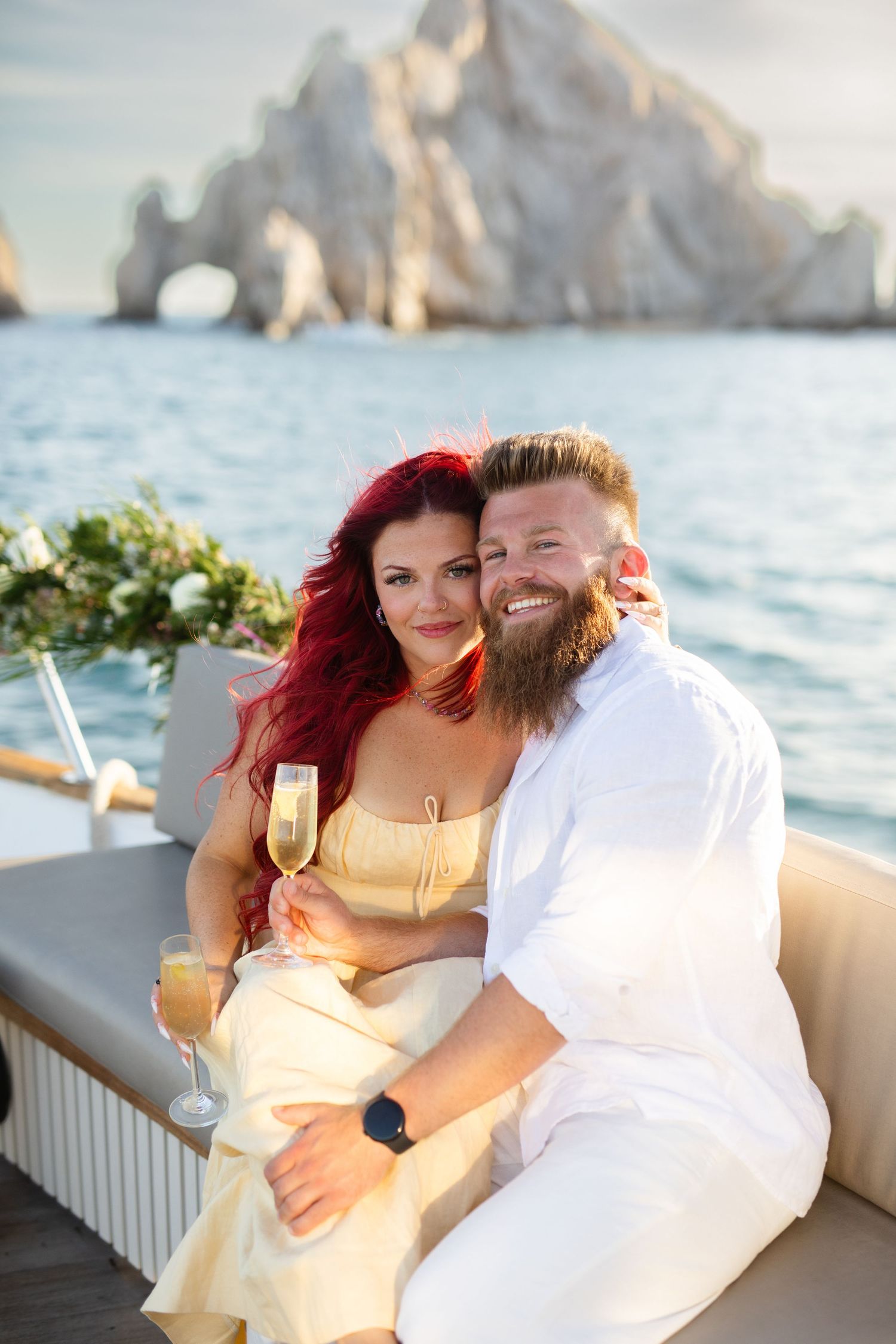 Smiling couple with champagne on a yacht near iconic rock arch in Cabo San Lucas at sunset.