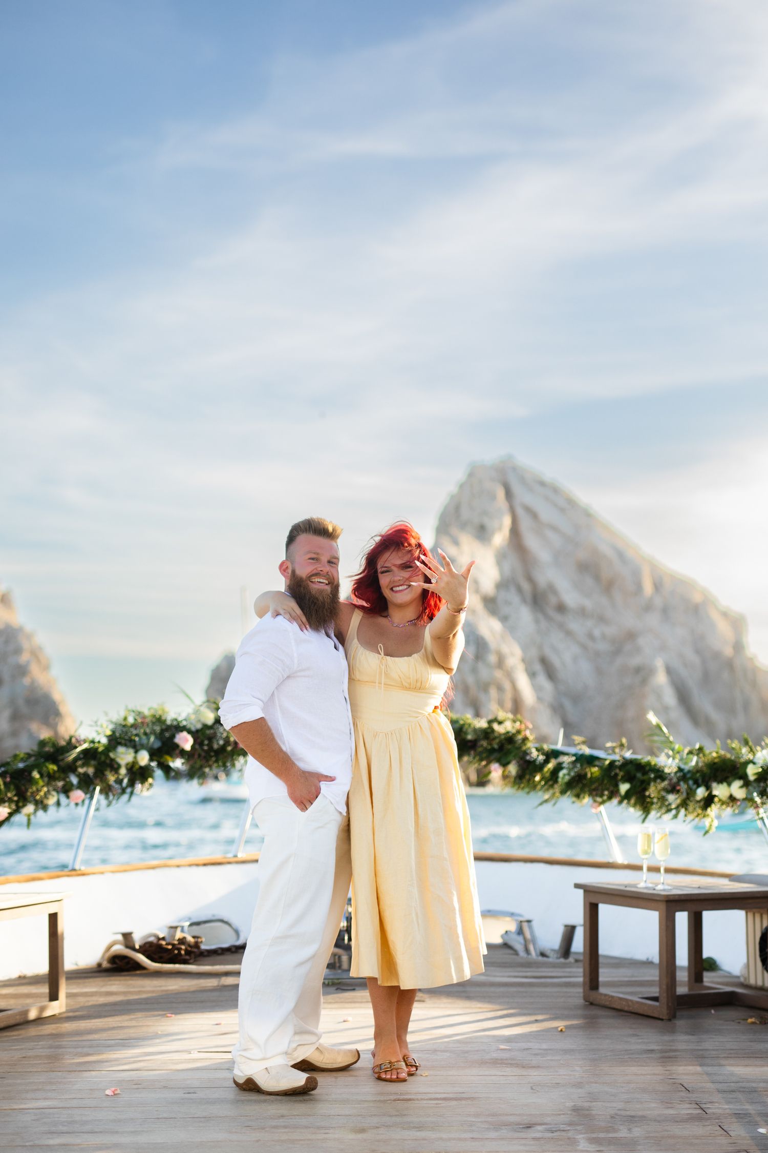 Couple posing on a wooden deck with ocean and rocky cliffs in background, woman in yellow dress, man in white outfit.