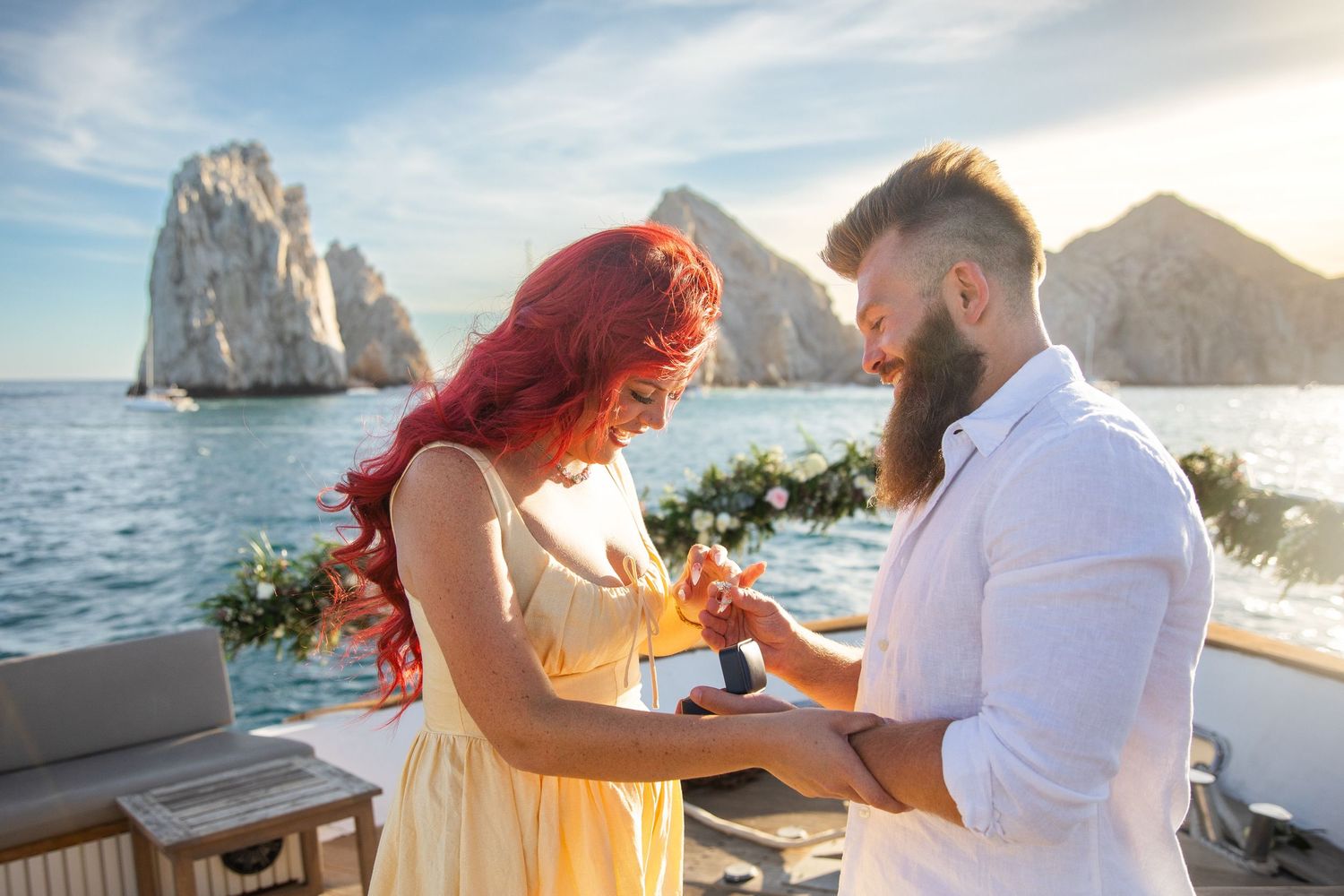 Couple sharing romantic proposal moment on yacht deck with stunning ocean rock formations at sunset.