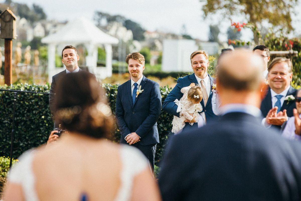 Groom smiles as bride approaches altar at outdoor wedding ceremony with white gazebo.